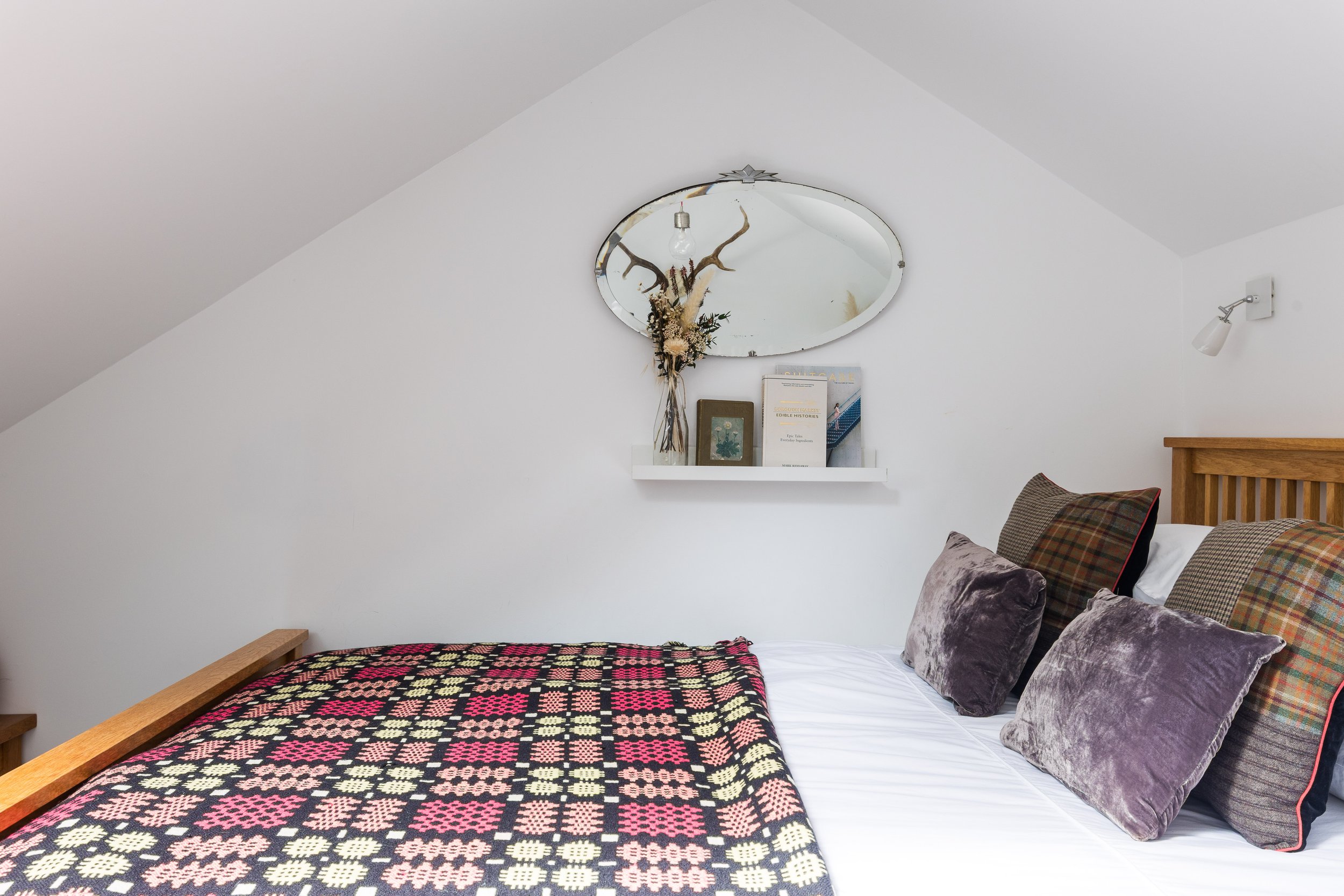 A bedroom with a white sloped ceiling, a bed with colorful patterned blanket, several pillows, a wall shelf with books and a vase with dried flowers, a mirror with antler decor, and a picture frame.