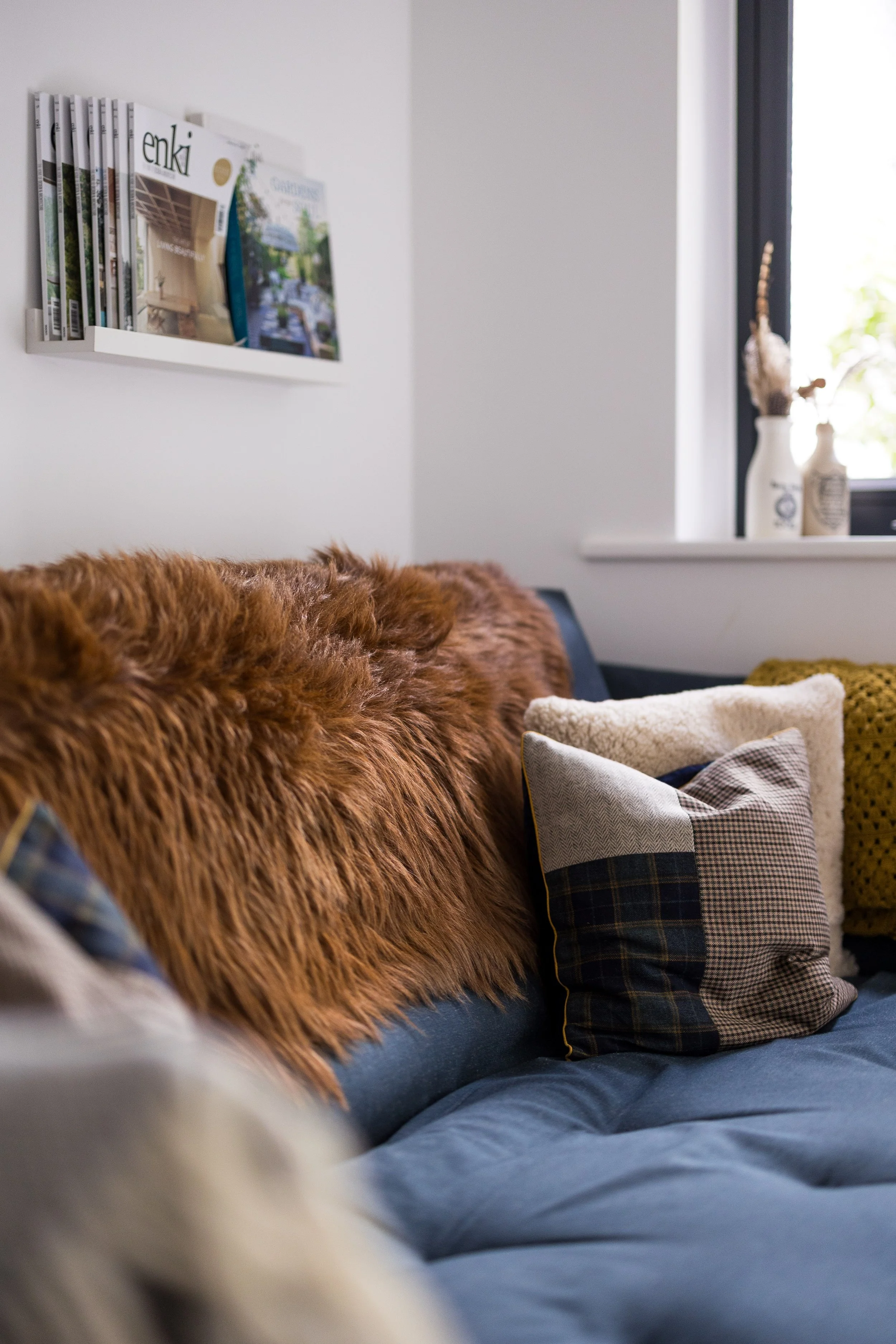 View of a cozy living room corner with a sofa covered in a blue slipcover, adorned with plaid and textured cushions, a fluffy rug, and a wall-mounted magazine rack with reading materials. A window with sunlight comes through, illuminating decorative vases in the background.