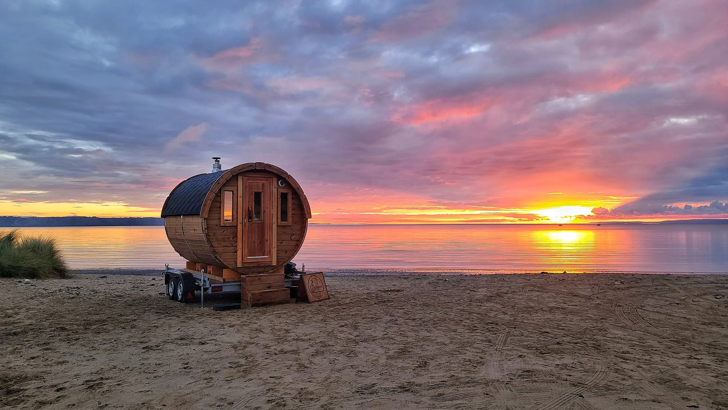 A small wooden barrel-shaped sauna on wheels parked on a sandy beach at sunset with colorful clouds in the sky and calm water in the background.