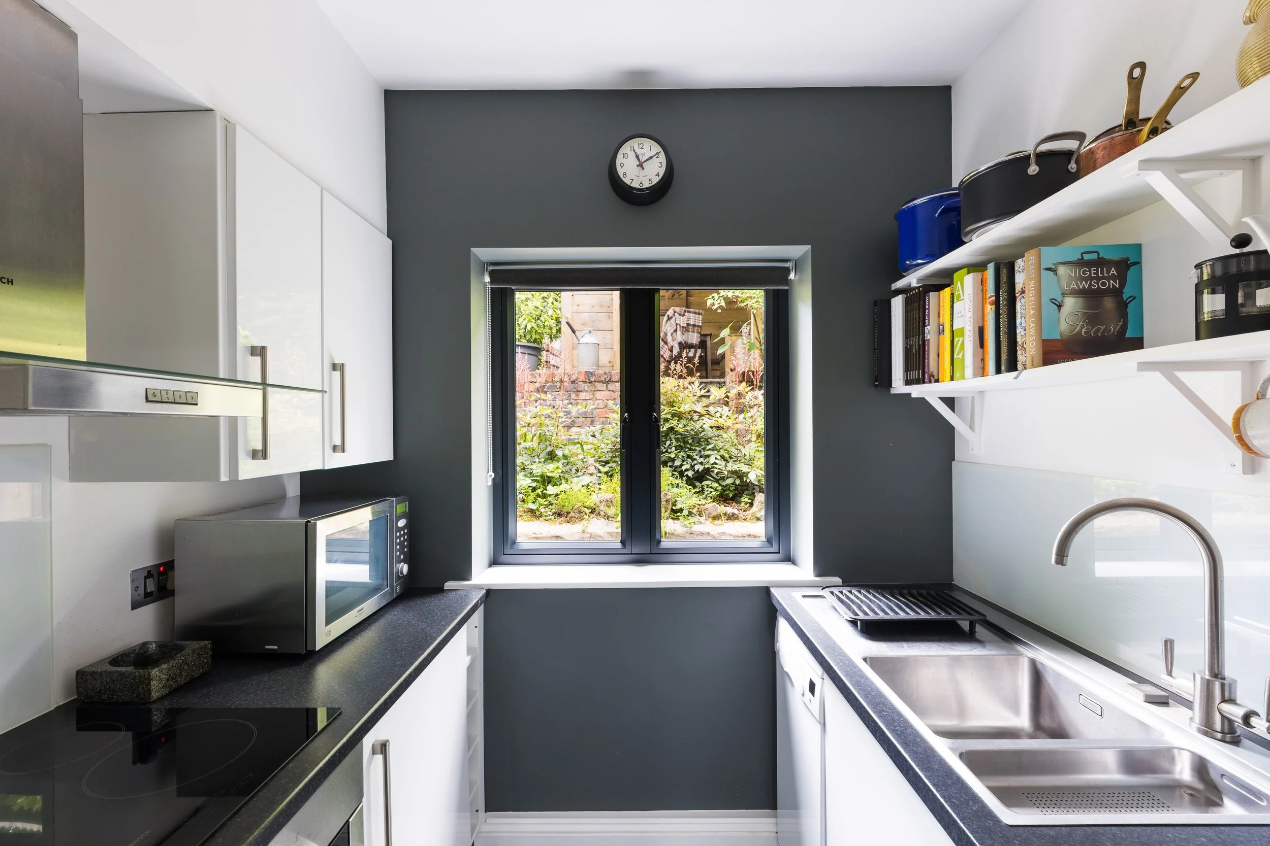 Modern kitchen with white cabinets, black countertops, a window overlooking a backyard, a clock on the dark gray wall, and various kitchen appliances and books on shelves.