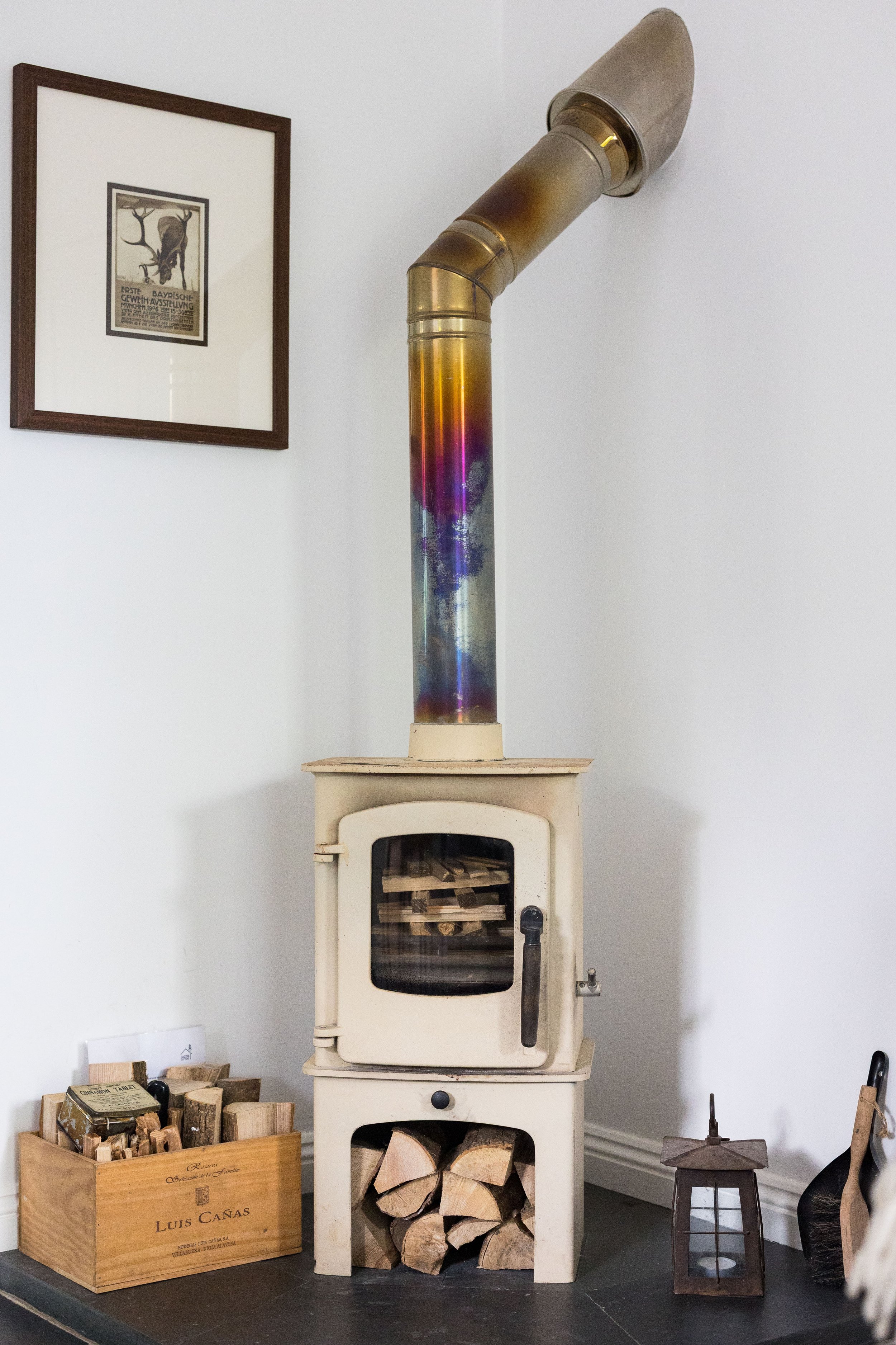A vintage beige stove with firewood stored below, a rainbow-colored metal chimney pipe above, a wooden box filled with chopped wood on the floor to the left, a lantern with a metal handle to the right, and a framed picture of a stag on the wall.