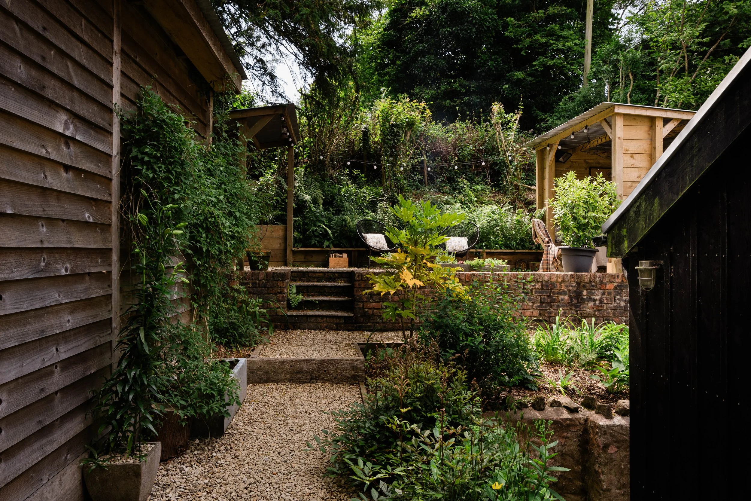 A lush backyard garden with wooden structures, green foliage, potted plants, and outdoor seating on a gravel path.