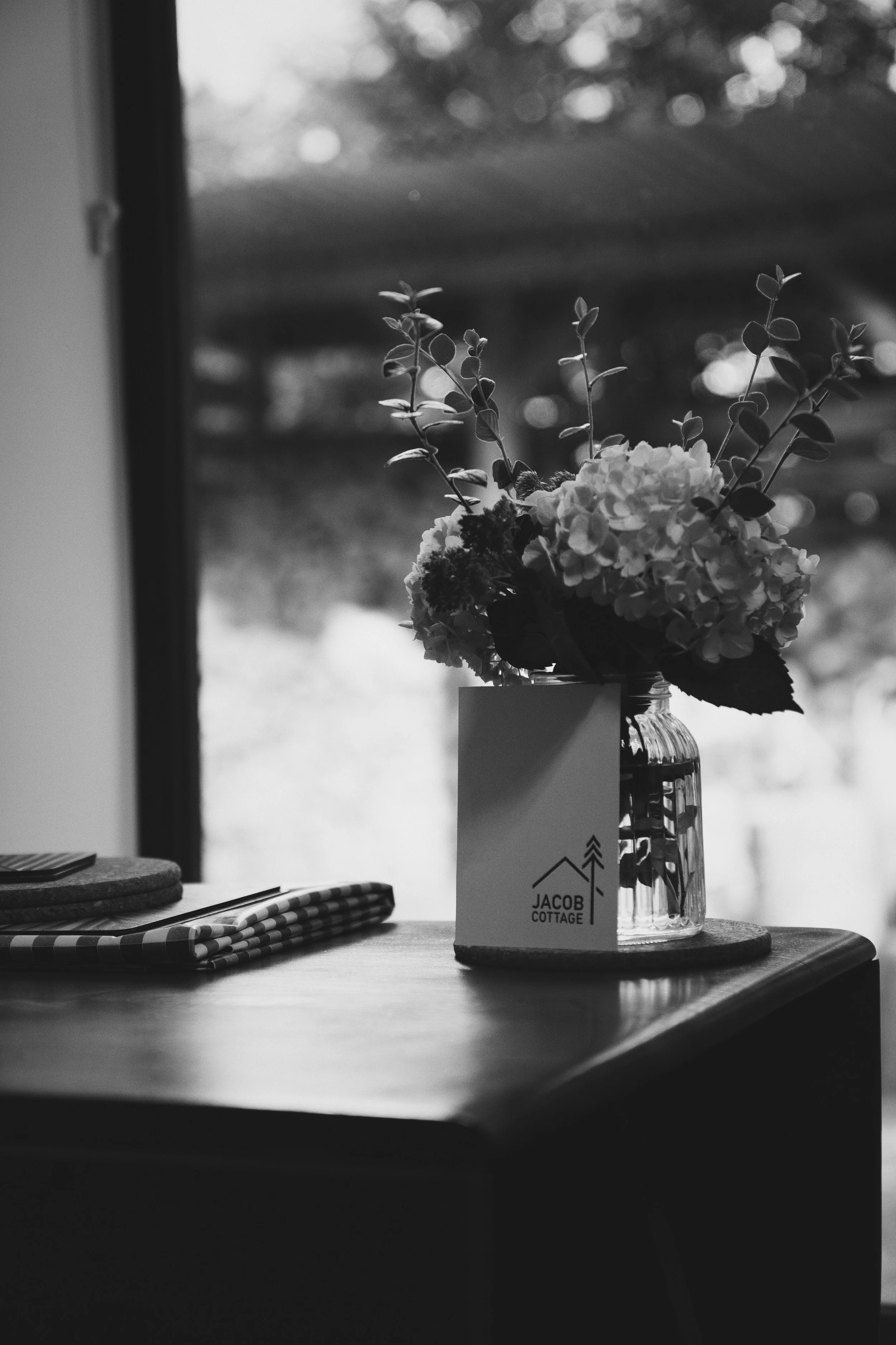 A table near a window holding a glass jar with flowers and a small card labeled "Jacob Cottage.