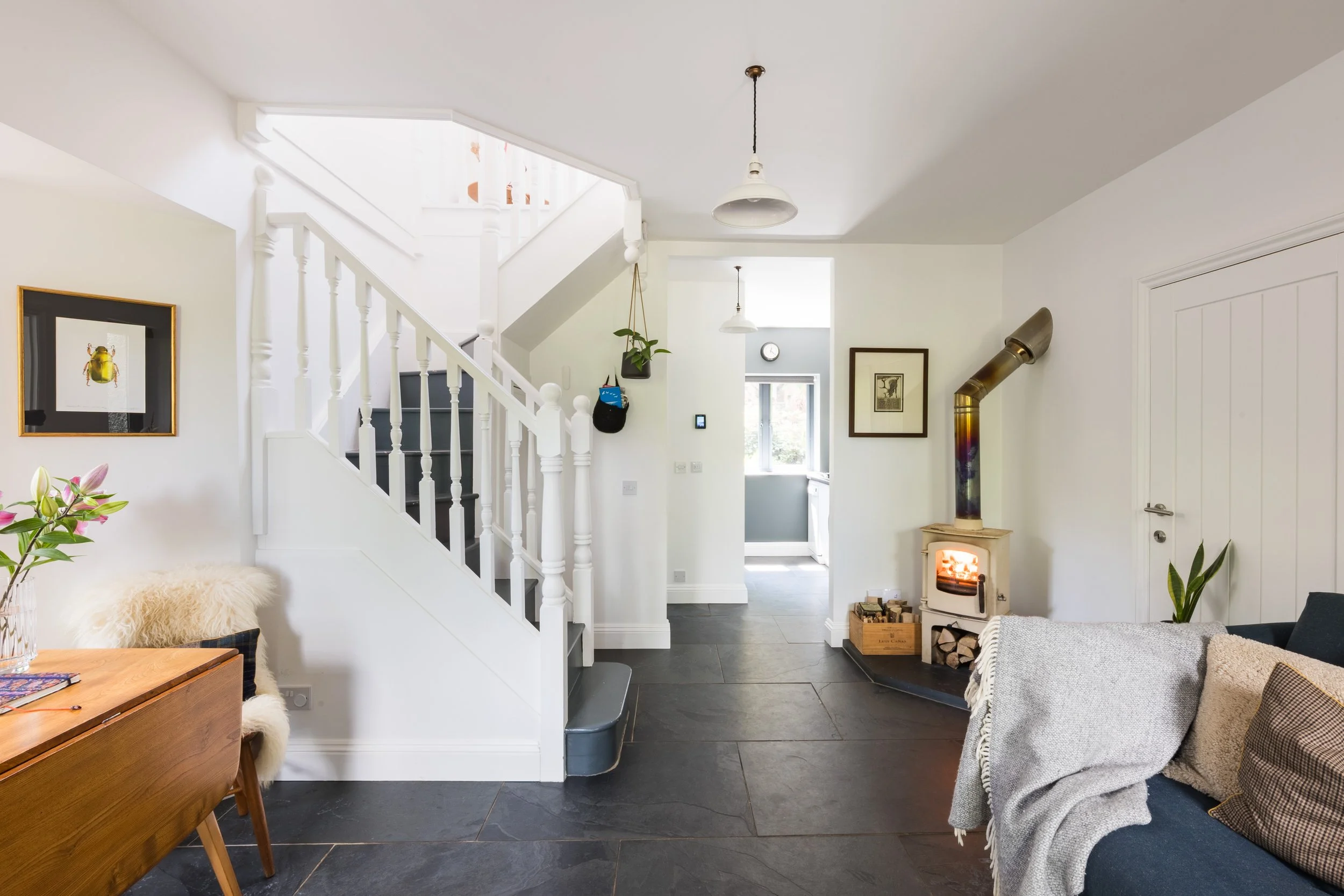 Living room with a staircase, hanging light fixtures, a wood stove, framed artwork, and furniture including a sofa and a wooden table with a sheepskin cover.