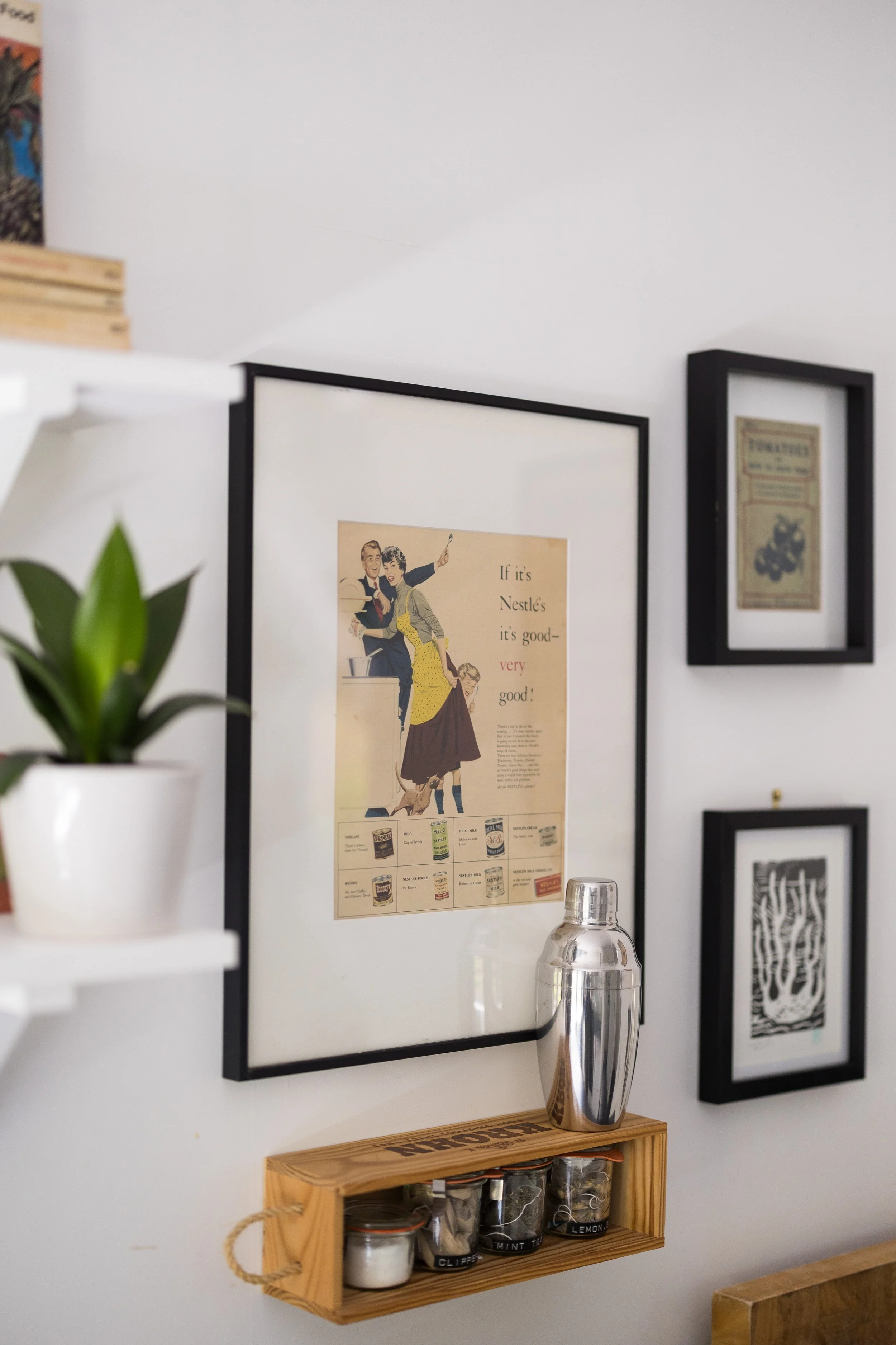 Decorative wall with framed vintage posters, a small wooden shelf with jars, a potted plant, and a metallic vase.