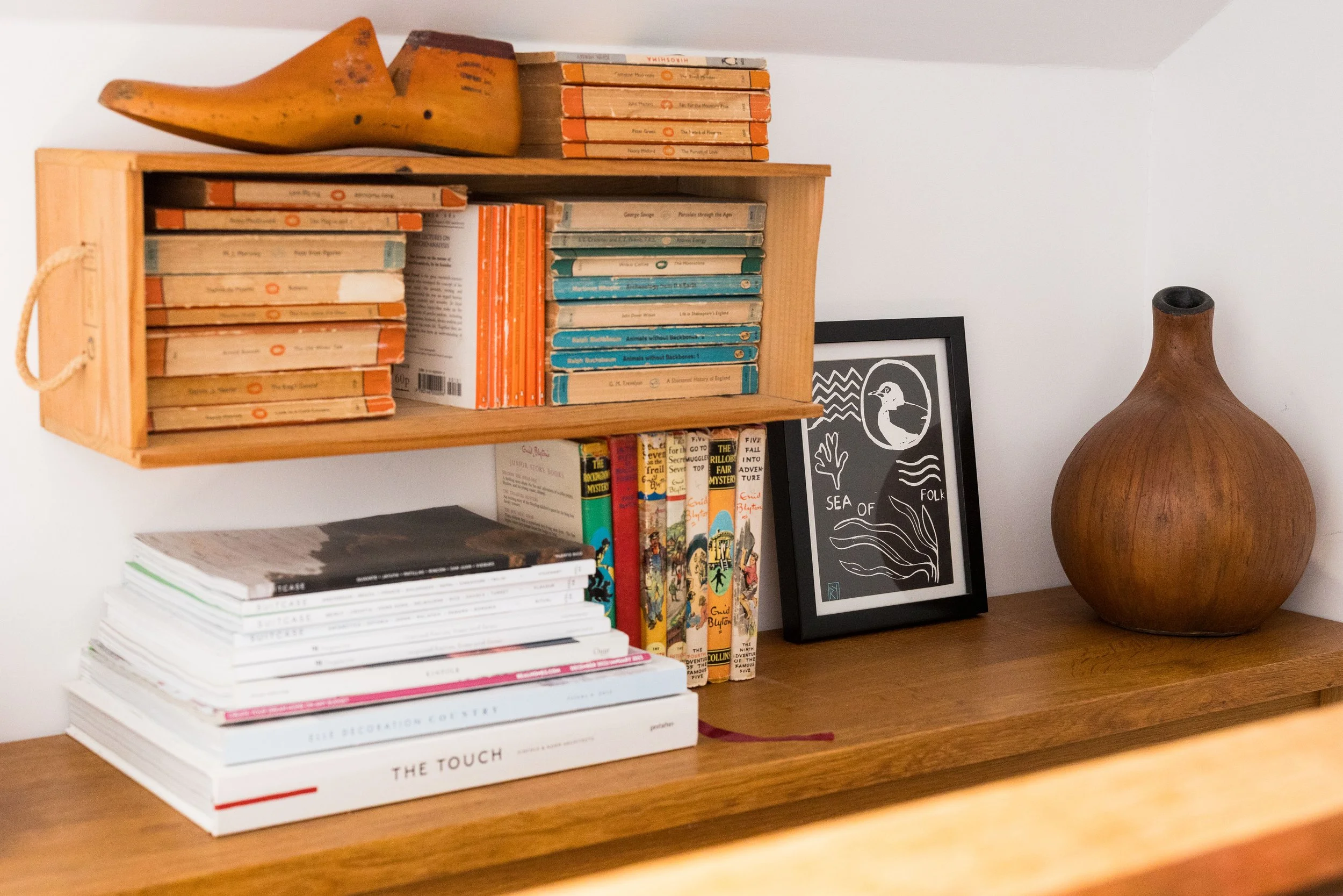 A wooden bookshelf with a decorative banana sculpture on top, multiple stacks of books including vintage orange-spined books, children's books with colorful covers, and a framed black-and-white print of a bird and seaweed, accompanied by a large brow