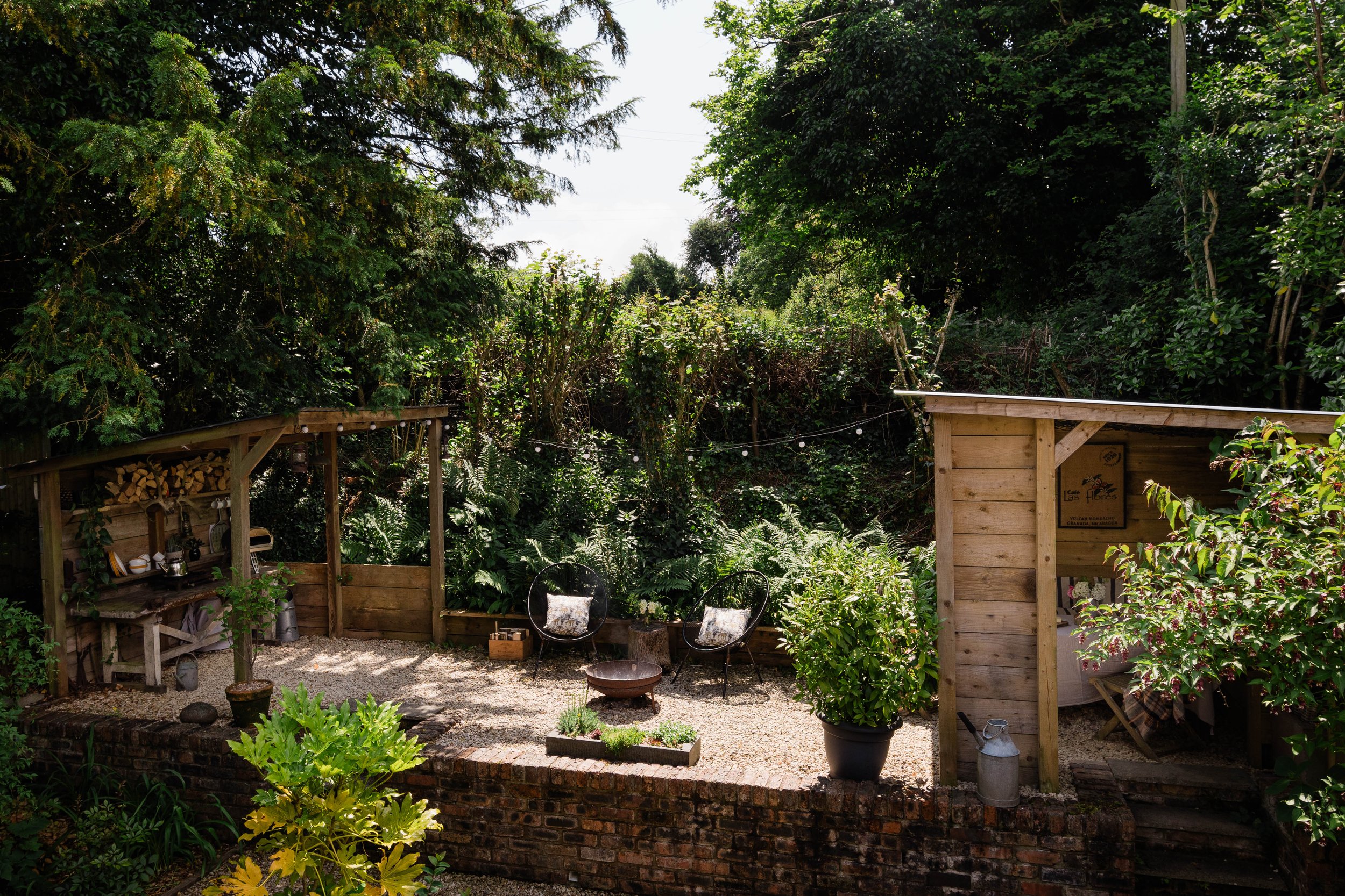 A cozy backyard garden with a wooden shed, black chairs with patterned cushions, potted plants, decorative items, and a backdrop of lush green trees and bushes.