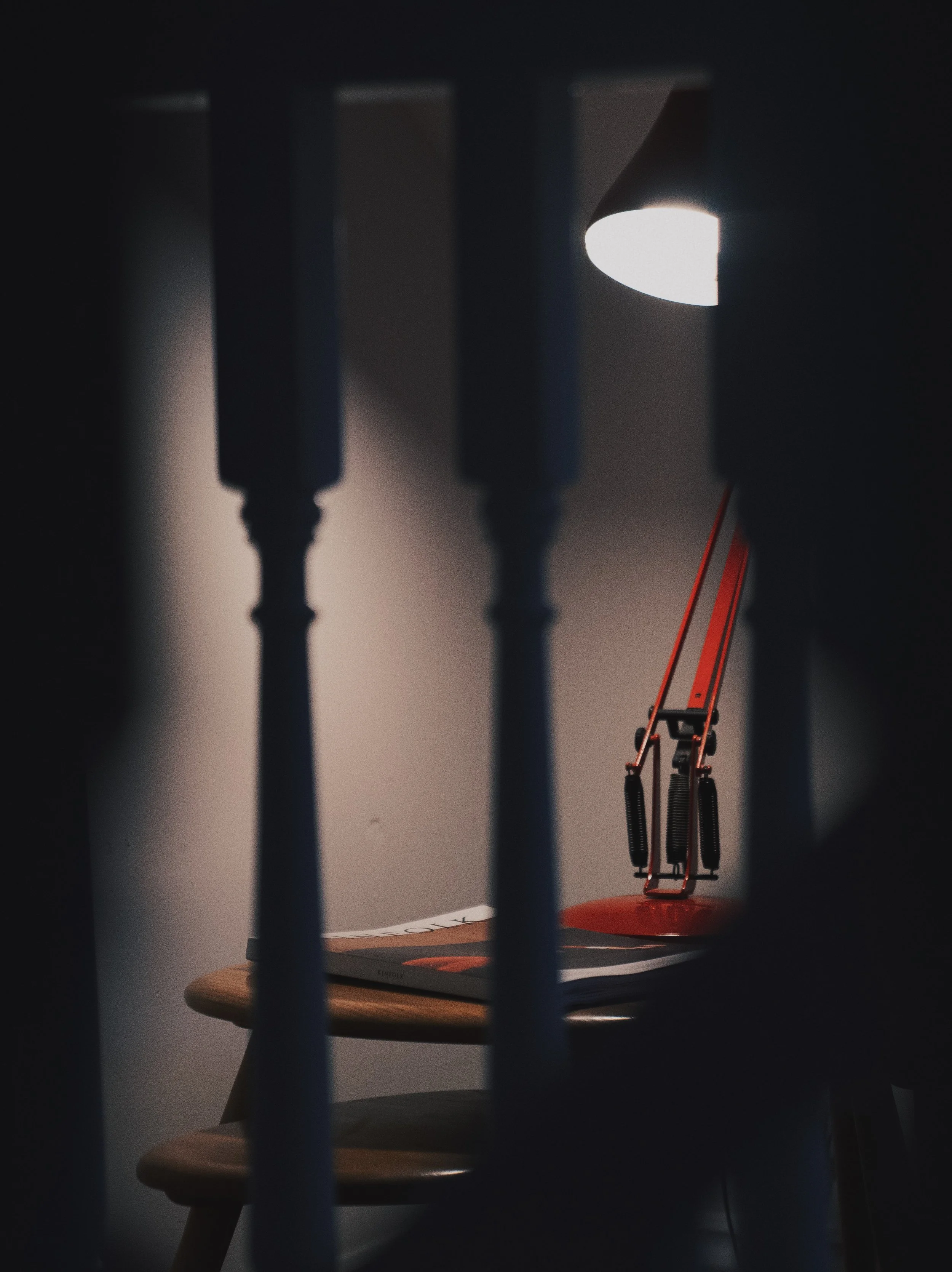 A small desk lamp shining bright on a wooden table, with a closed book nearby, it is viewed through the cracks of a blue rail or similar object.