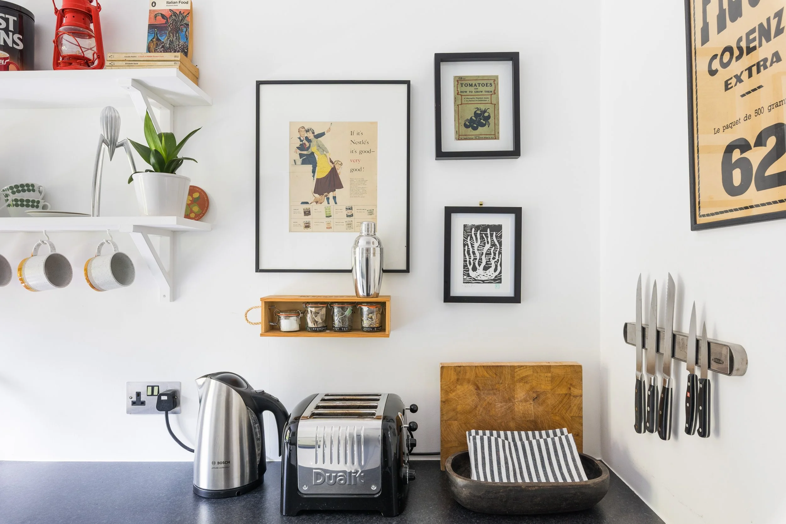 A kitchen countertop with a silver toaster, a coffee machine, a cutting board, a knife holder with knives, and a black sink. On the wall, there are framed posters and artwork, a plant in a white pot, hanging mugs, and a shelf with books, a red lanter