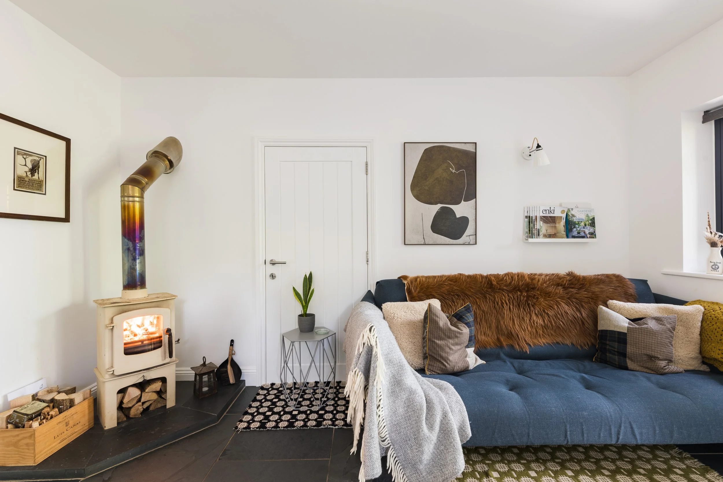 Cozy living room with a wood-burning stove, abstract art on the white walls, a dark blue sofa with various pillows and throws, a small side table with a plant, and a window letting in natural light.