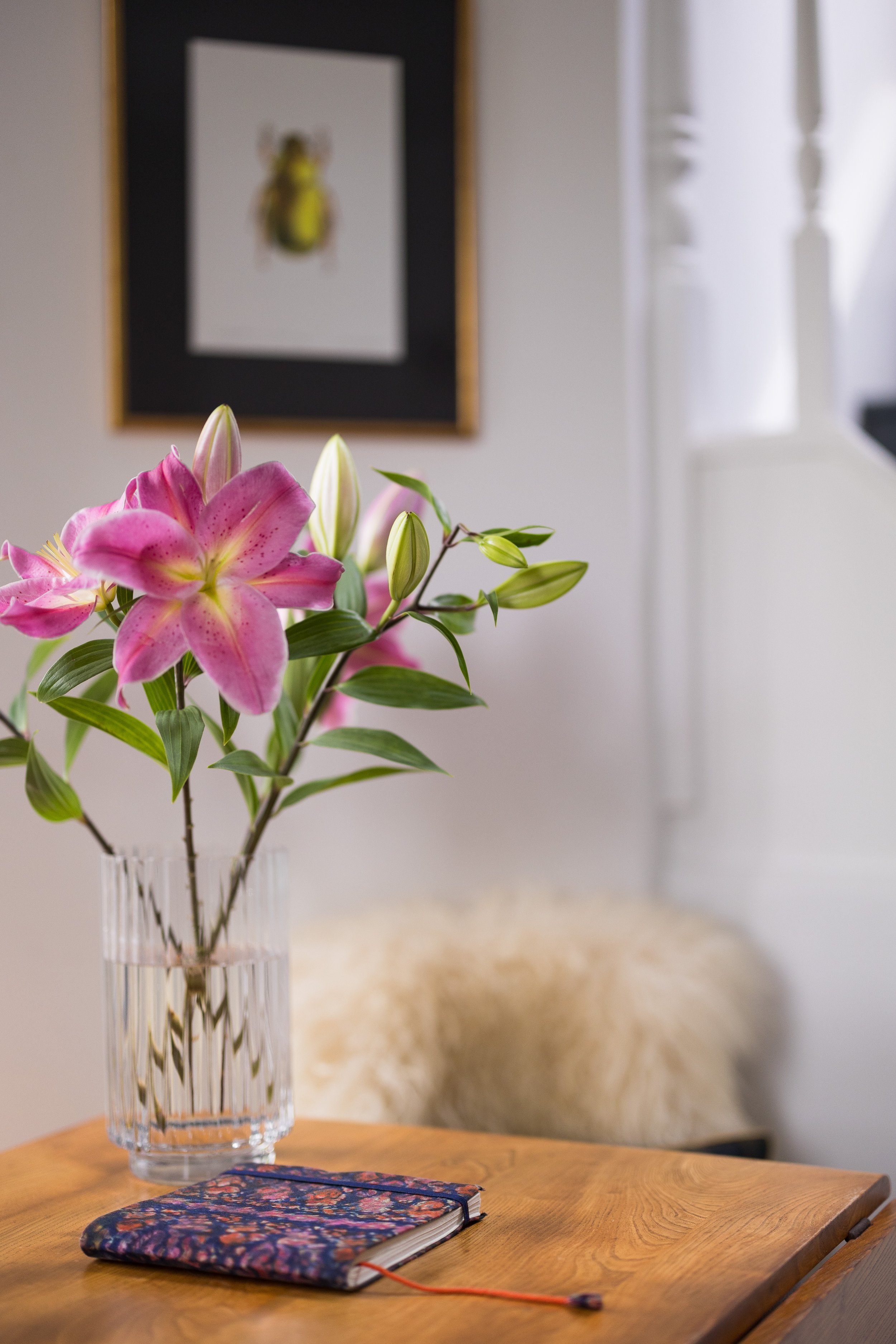 A pink lily flower with several buds arranged in a clear glass vase on a wooden table. A colorful floral-patterned notebook with a red elastic band and a pen are also on the table. In the background, a framed picture of a beetle is hanging on a white