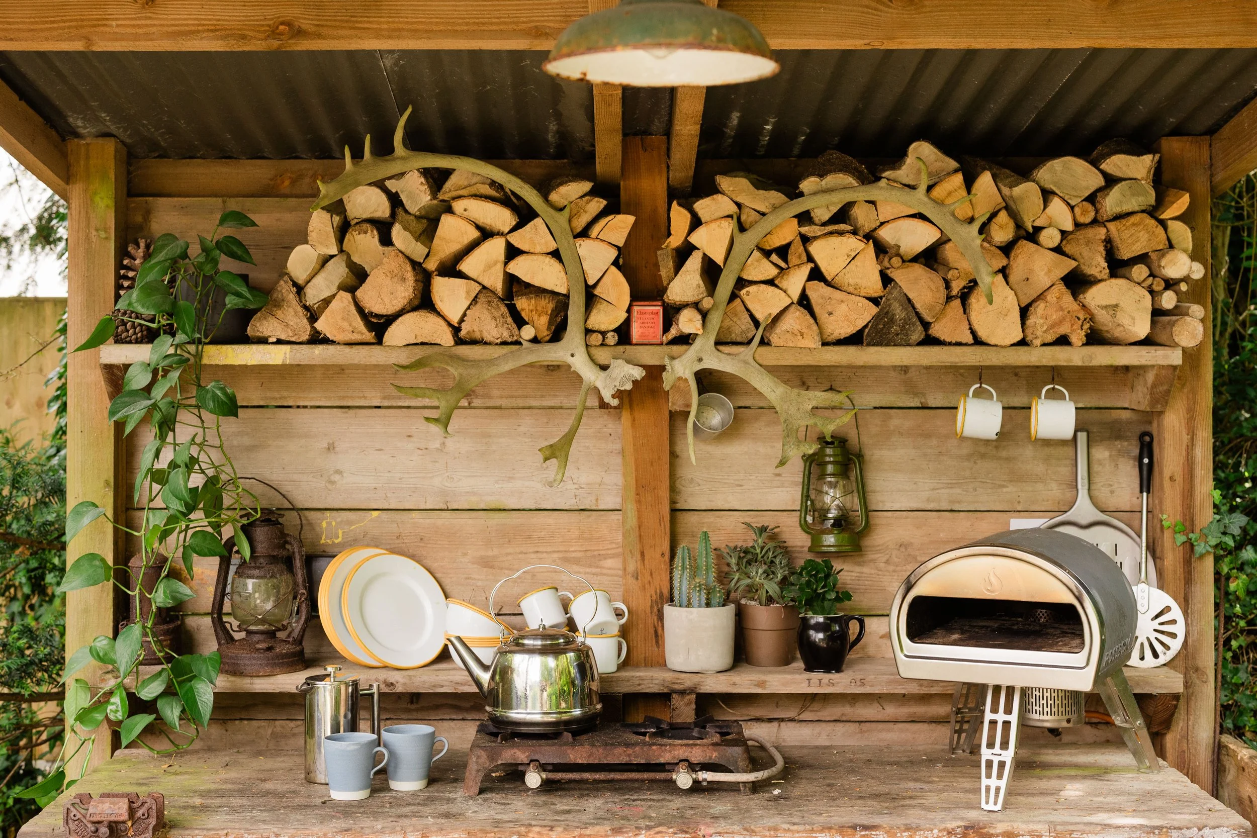 Wooden outdoor kitchen with stacked firewood, kitchen utensils, cups, a kettle, a toaster, and potted plants, under a corrugated metal roof.