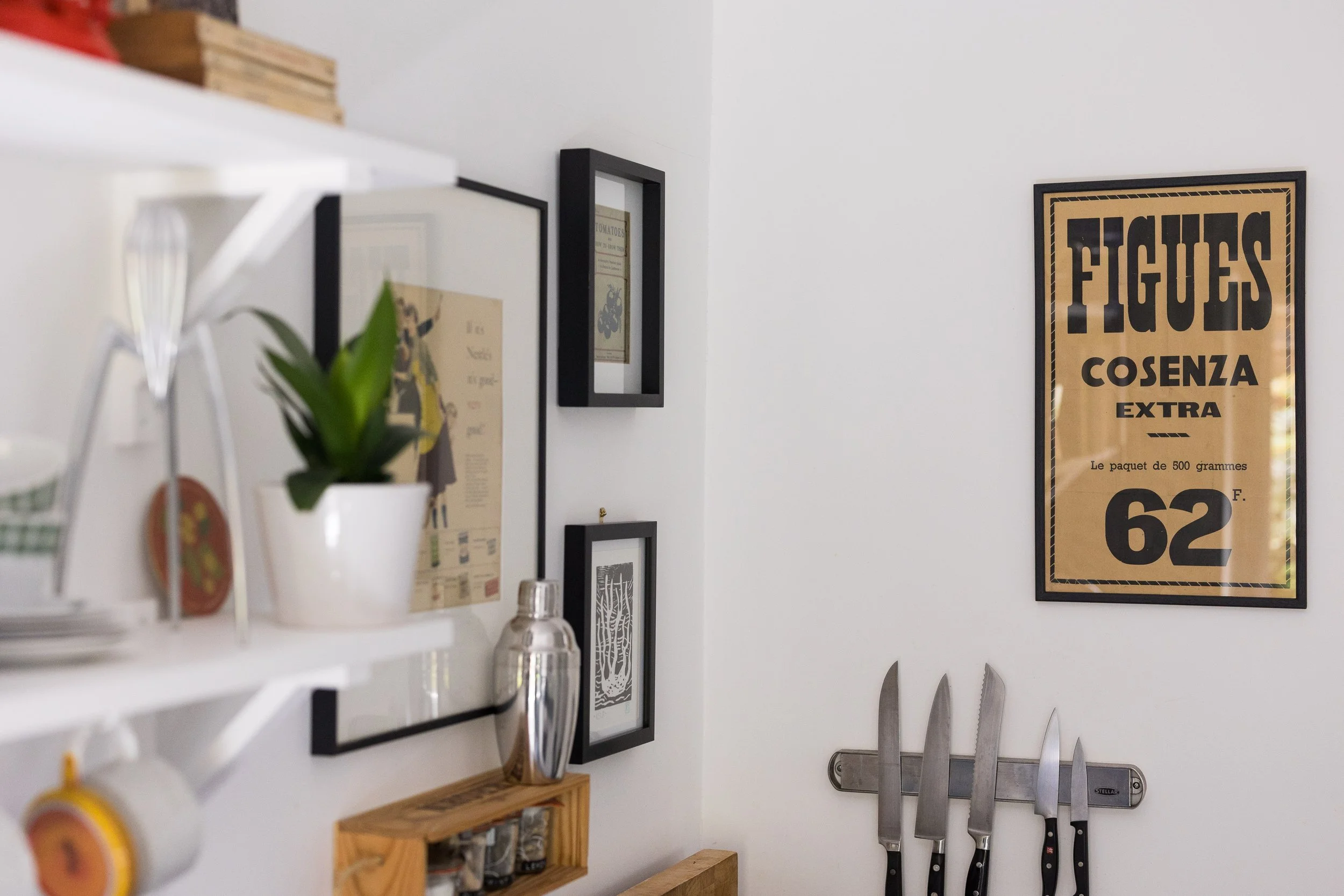 A kitchen with kitchen knives mounted on a wall, framed wall art, and a shelf with plants and kitchen items.