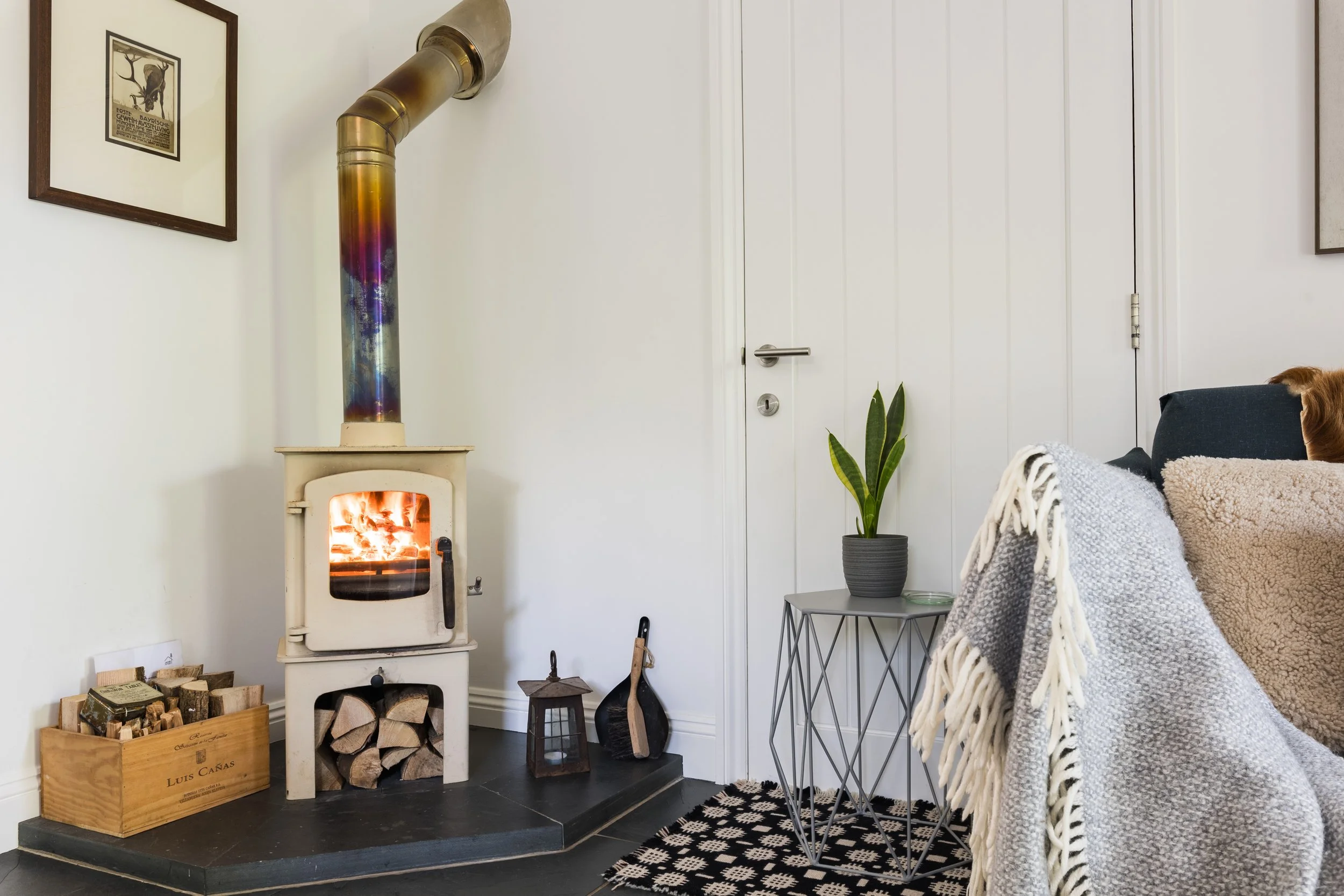 Cozy living room corner featuring a wood stove with a chimney pipe, logs in a wooden crate, a potted snake plant, a small table, and chairs with blankets, alongside framed artwork on the white wall.