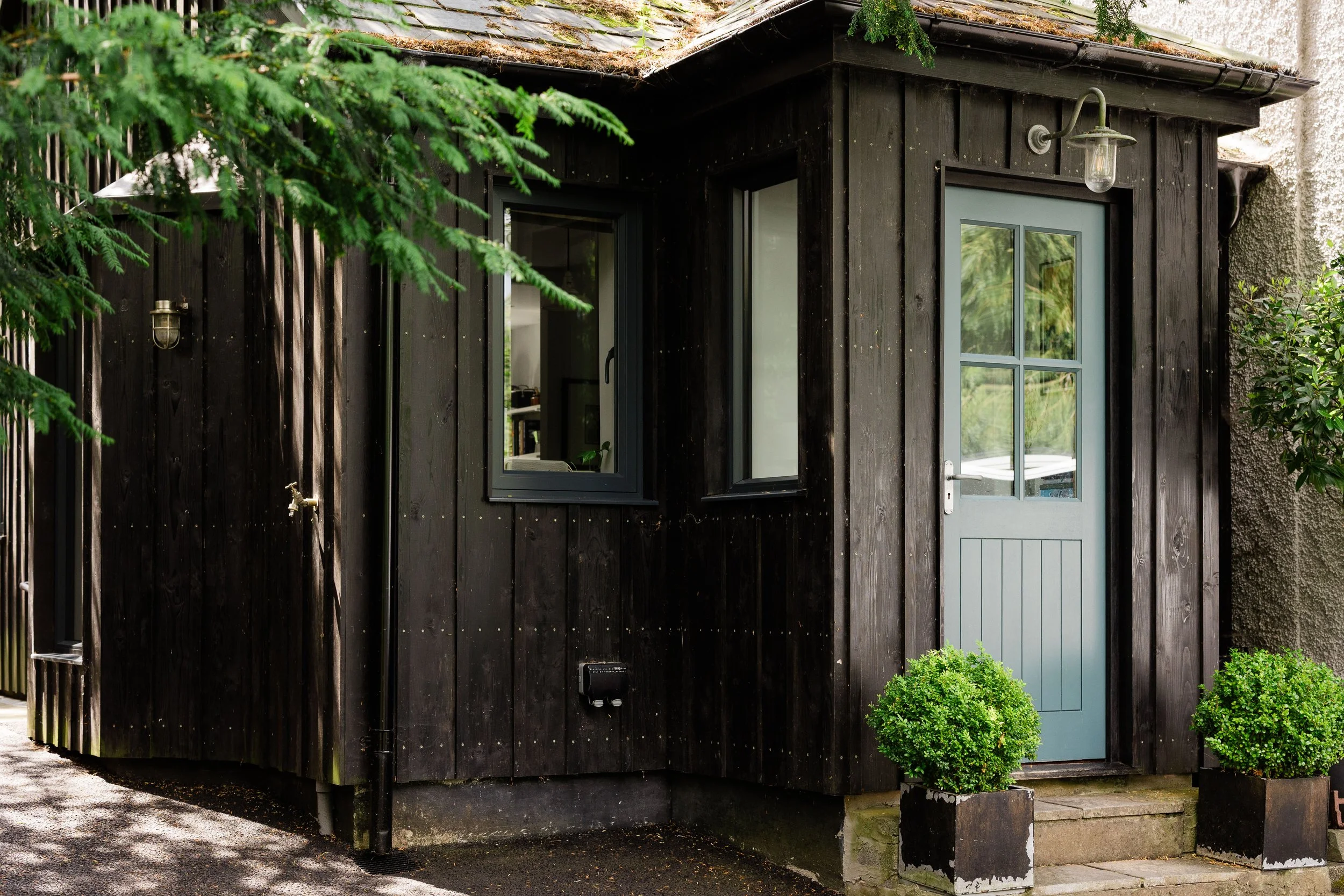 A black wooden exterior of a small cottage or shed with a gray door, two windows, and outdoor lights, surrounded by greenery and trees.