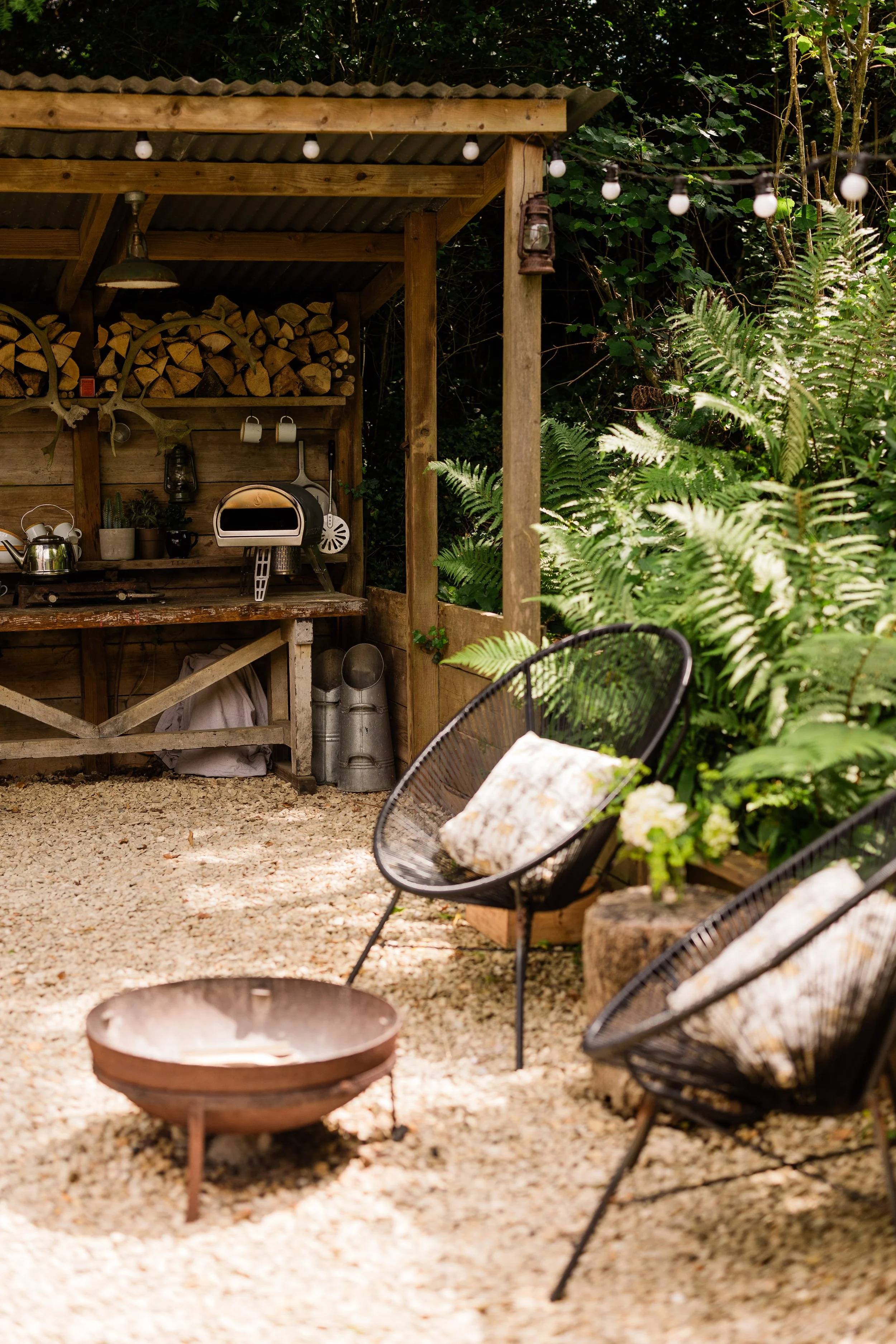 An outdoor rustic kitchen area with a wood firewood rack, a small oven, and cozy chairs with cushions, surrounded by lush green plants and string lights.