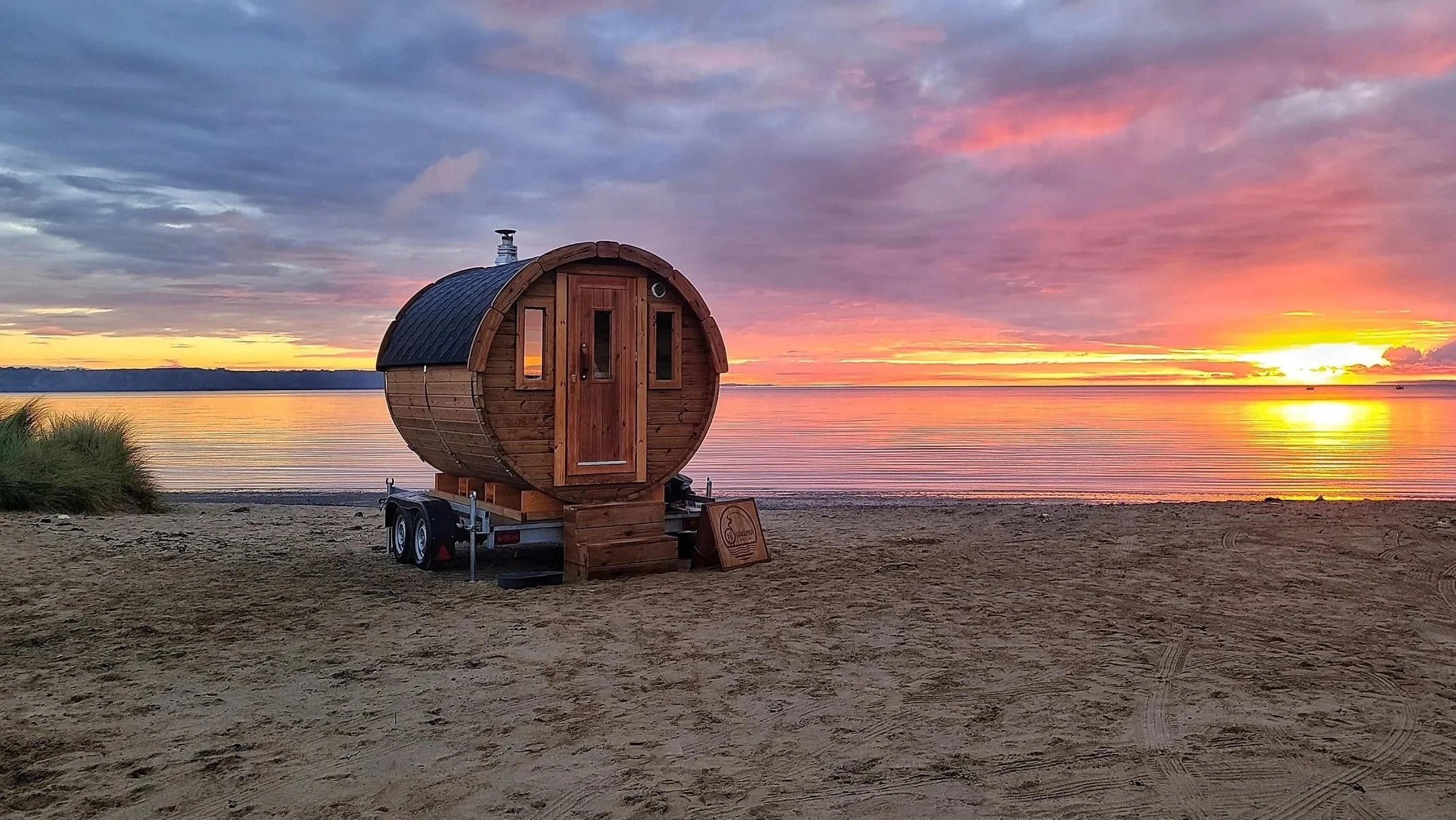 A small wooden sauna on wheels located on a sandy beach at sunset, with colorful sky and calm water in the background.
