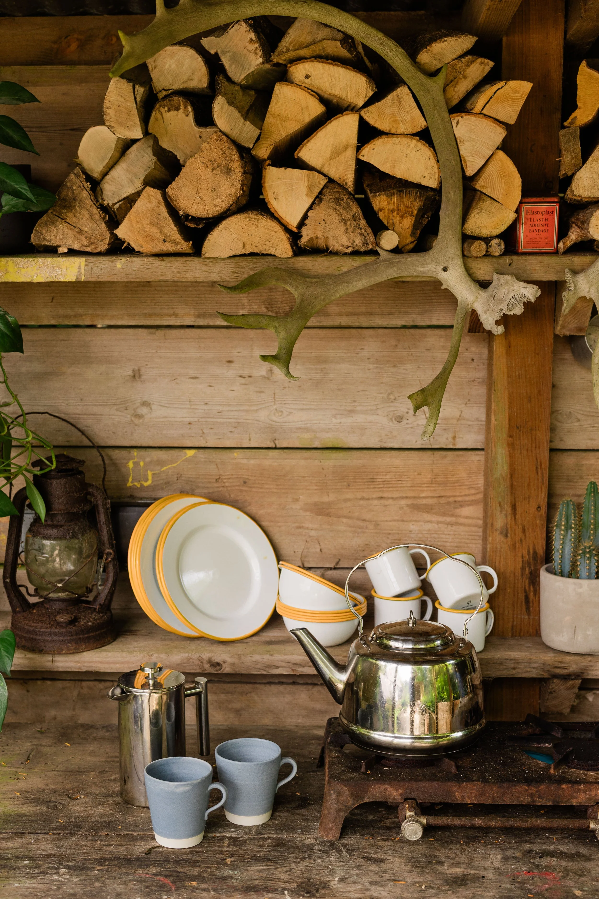 A wooden outdoor shelf with chopped firewood and a deer antler on top, a small orange box of Elastoplast bandages, a potted cactus, a glass plant bottle, yellow and white plates, cups, a teapot, a large stainless steel kettle, and a coffee pot on a rustic wooden table.