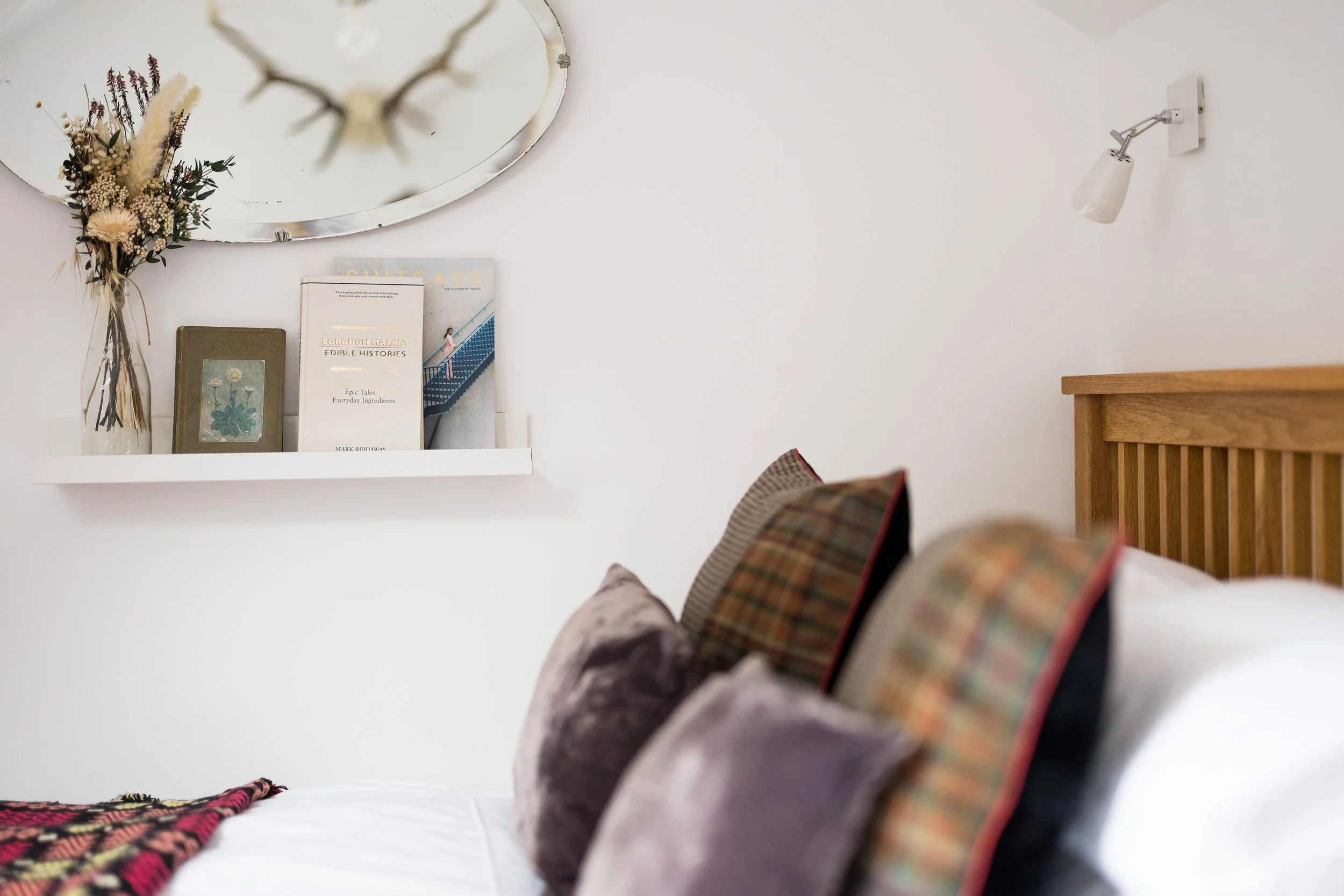 A bedroom corner with a white shelf holding books and a framed floral picture, a dried flower arrangement in a clear vase, and a wall-mounted reading lamp. Part of a bed with a plaid pillow is visible.