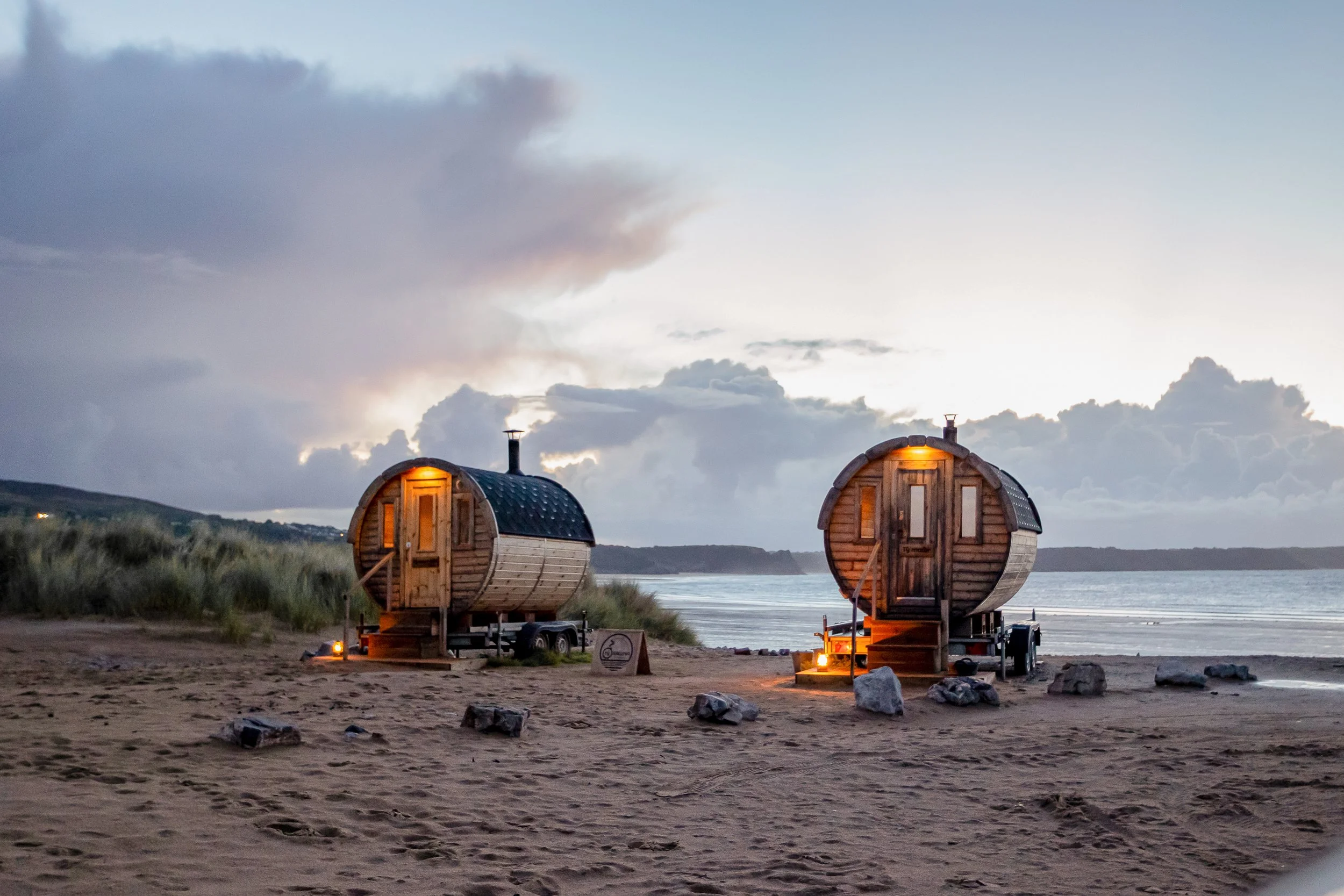 Two wooden barrel-shaped sauna cabins on trailers at the beach during dusk, with cloudy sky and water in the background.