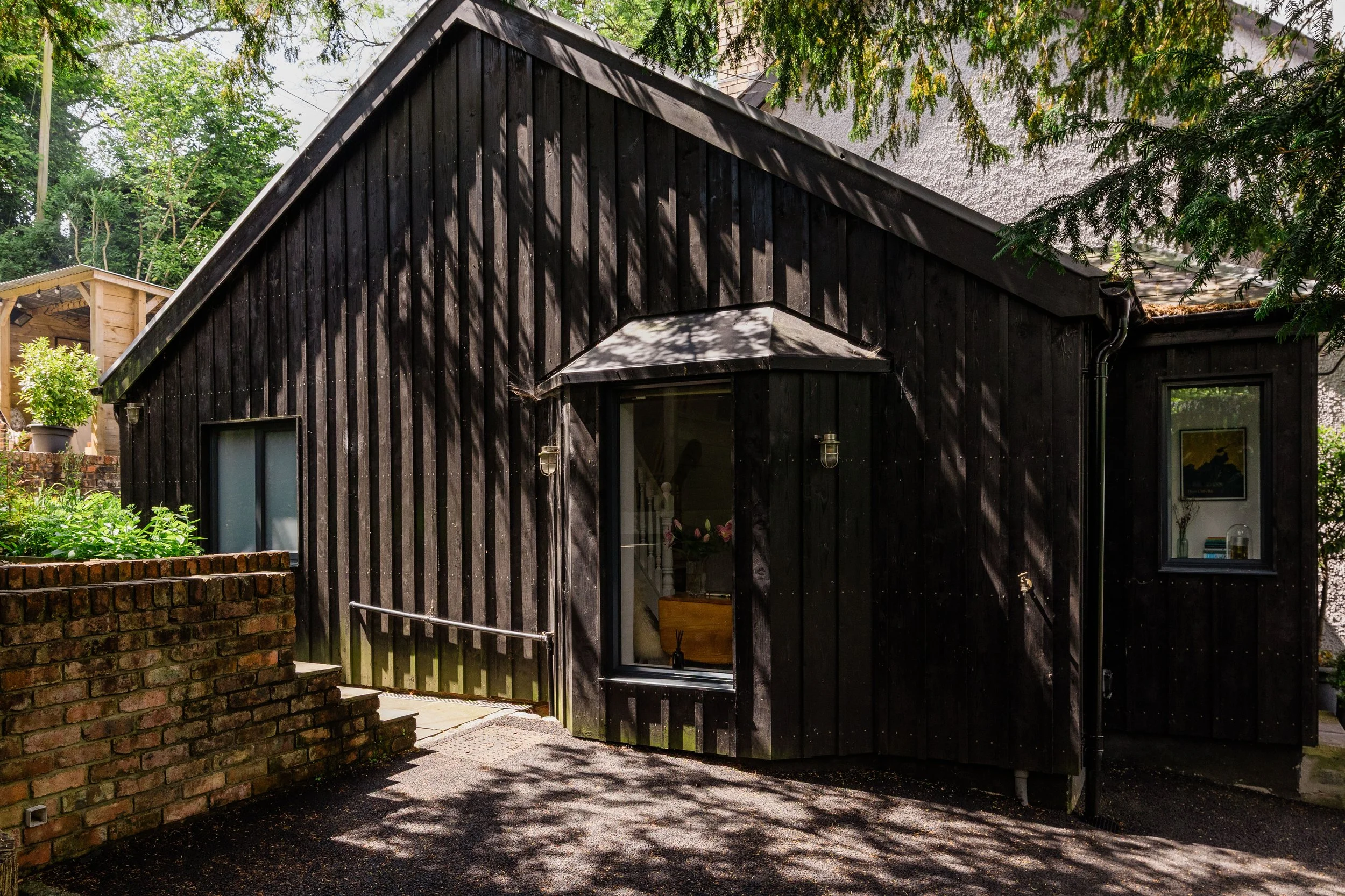 A black wooden house with large windows and a sloped roof, surrounded by greenery and brick steps leading to the entrance.
