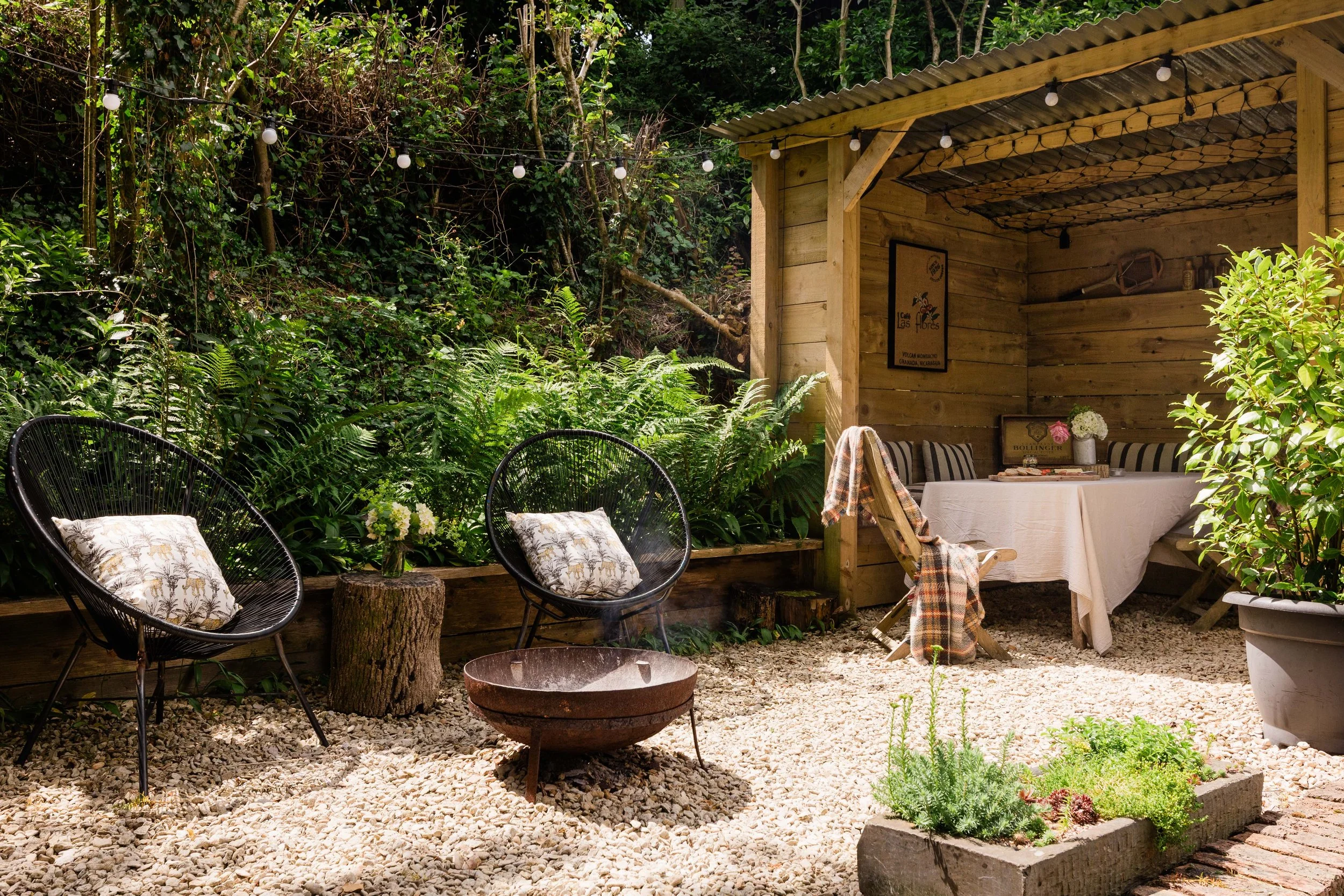 A cozy outdoor seating area with black wire chairs, pillows, and a round fire pit on a gravel surface, surrounded by lush green plants and trees. There is a wooden shed with string lights, a table with a tablecloth, and garden plants nearby.