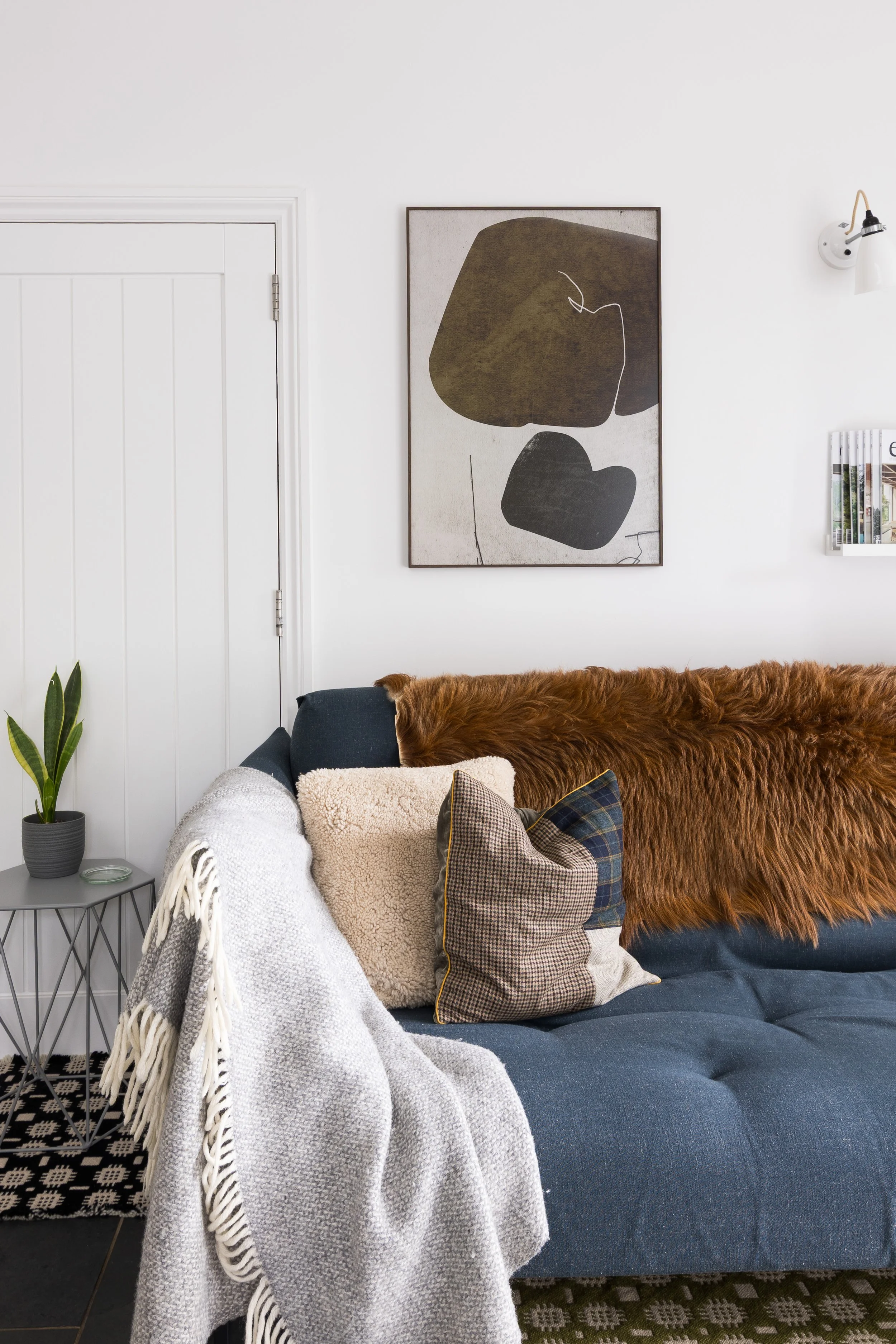 A living room with a white wall, minimalist abstract art, a dark blue sofa with assorted blankets and pillows, a small metal side table with a potted plant and candle, a brown furry throw, and a black and white geometric rug.