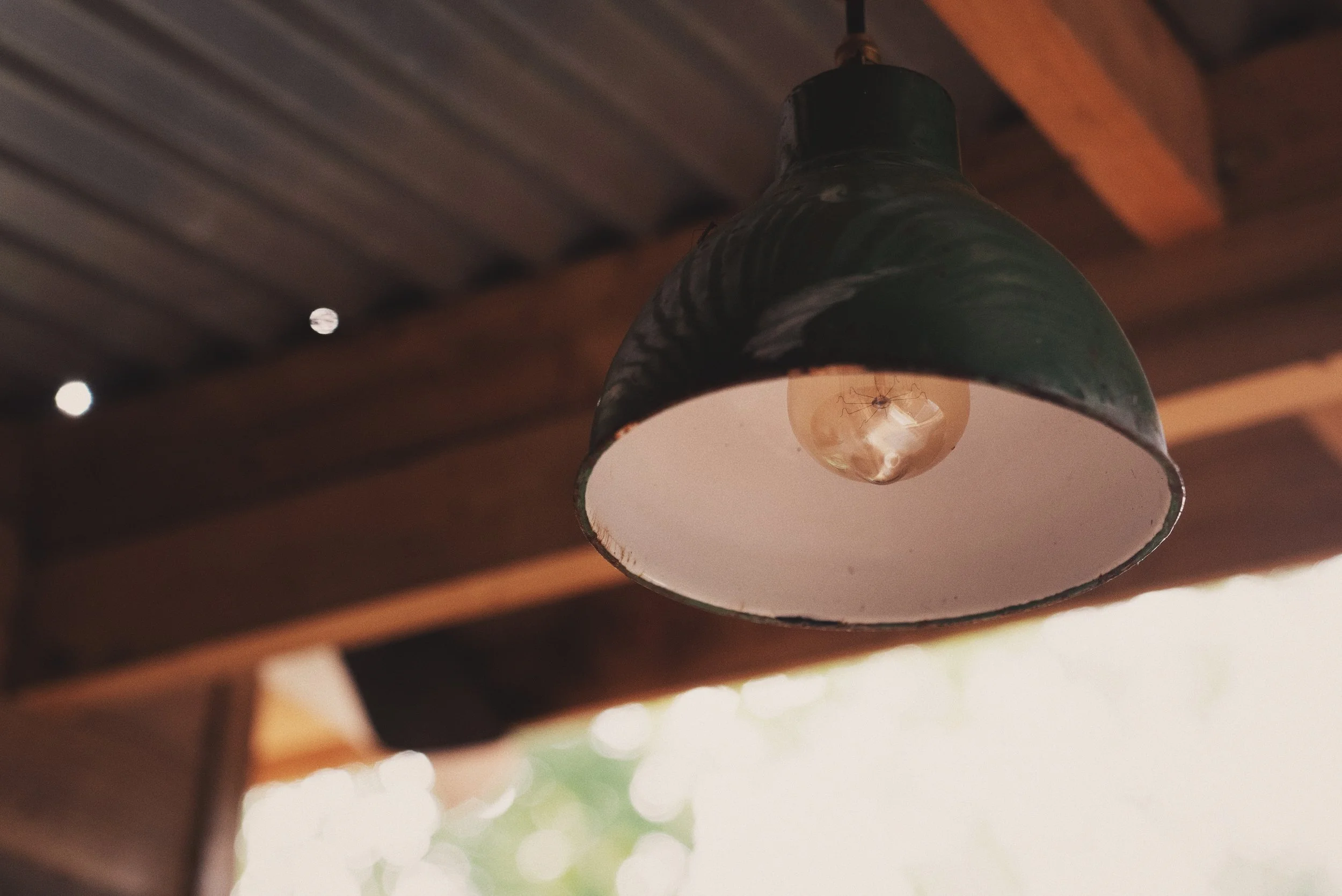 Close-up of an aged, rustic green metal pendant light hanging from a ceiling with wooden beams, with a spider hanging upside down inside the lampshade.