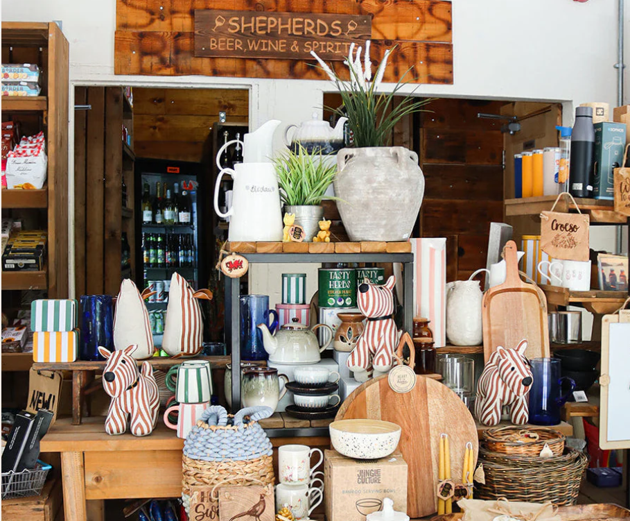 Display of various homeware items including ceramic zebra-shaped figurines, mugs, vases, bowls, wooden cutting boards, and plants on a rustic wooden shelf at Shepherds store, which sells beer, wine, and spirits.