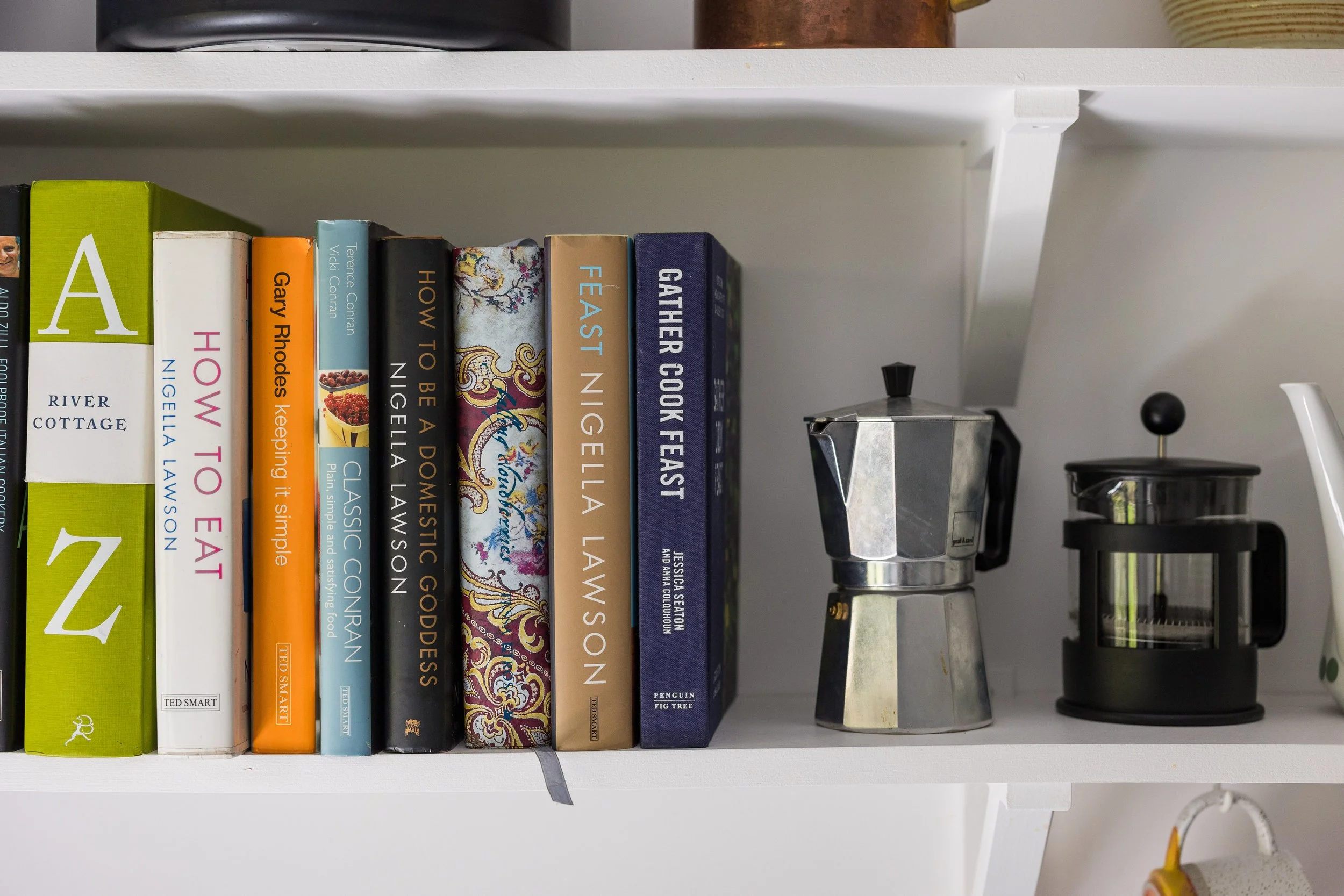 Close-up of a white bookshelf displaying a row of colorful books, a moka pot, and a small black pot.