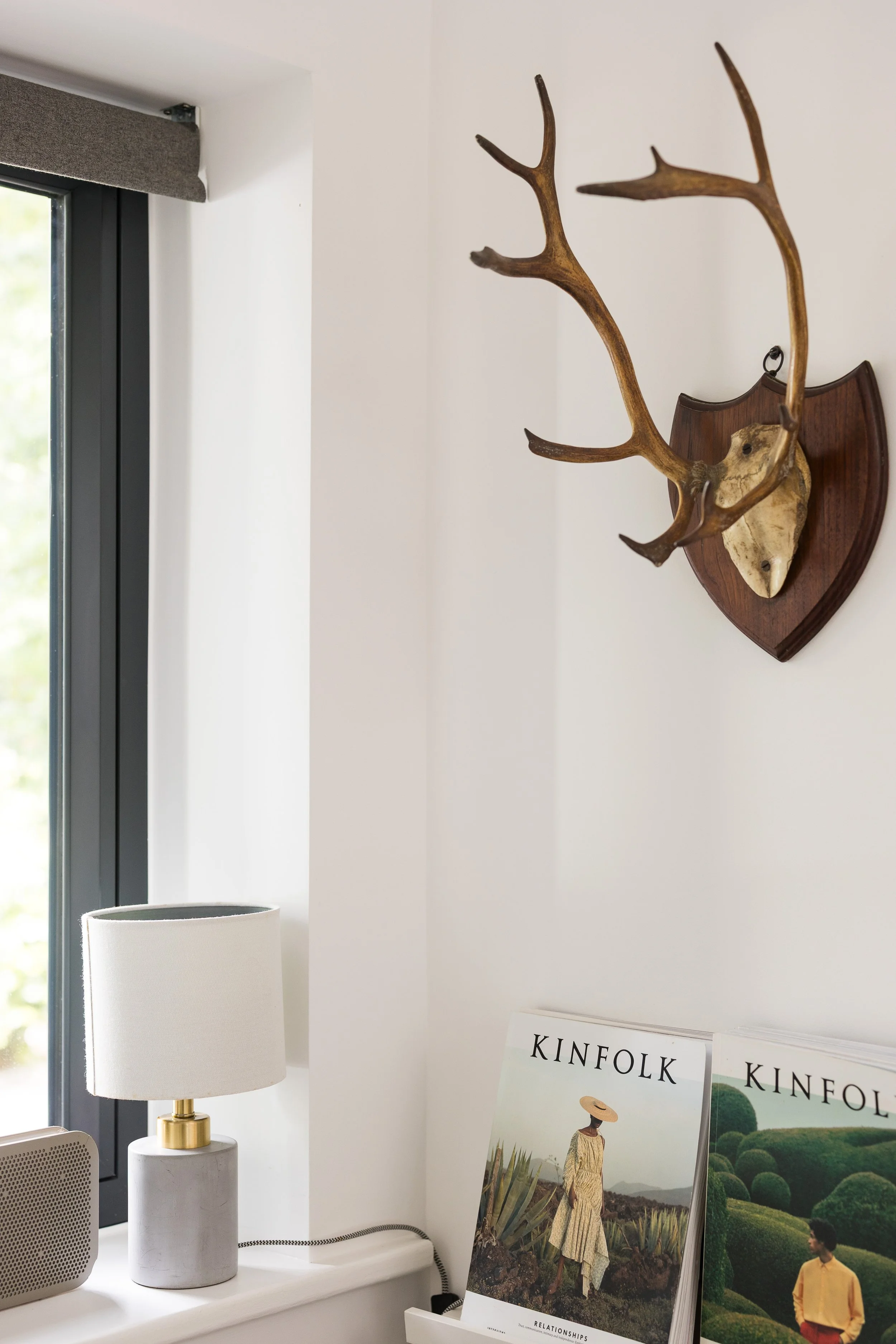 Interior of a room with a window, a white lamp with a gold accent, a speaker, and a magazine rack with Kinfolk magazines. A decorative mounted deer head with antlers is on the wall.