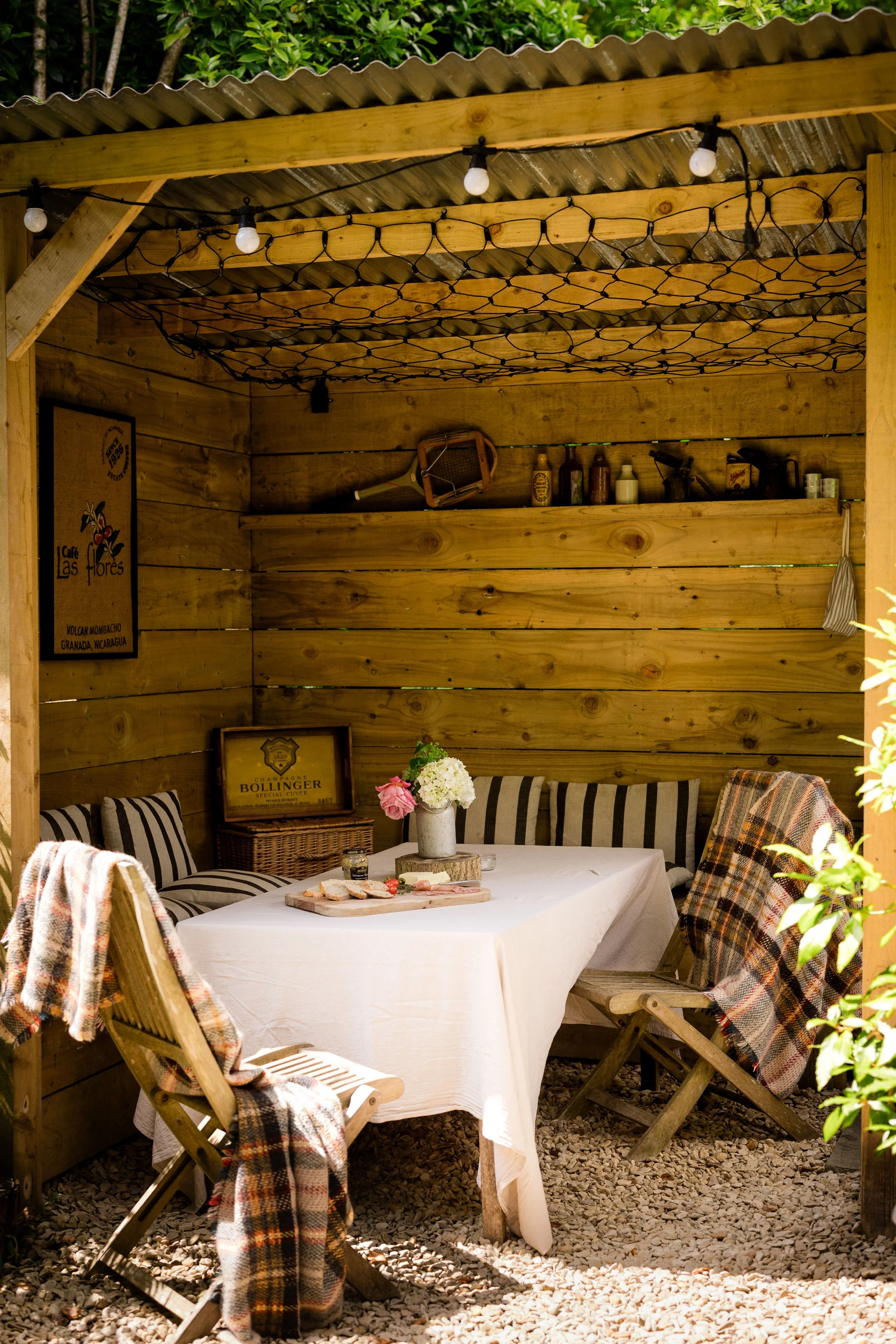Outdoor wooden patio with a white tablecloth-covered table set for a meal, surrounded by wooden chairs with plaid blankets. A vase with flowers and a wooden cutting board with food are on the table. The patio is enclosed by wooden walls with shelves holding bottles and decorative items, under a corrugated metal roof with string lights.