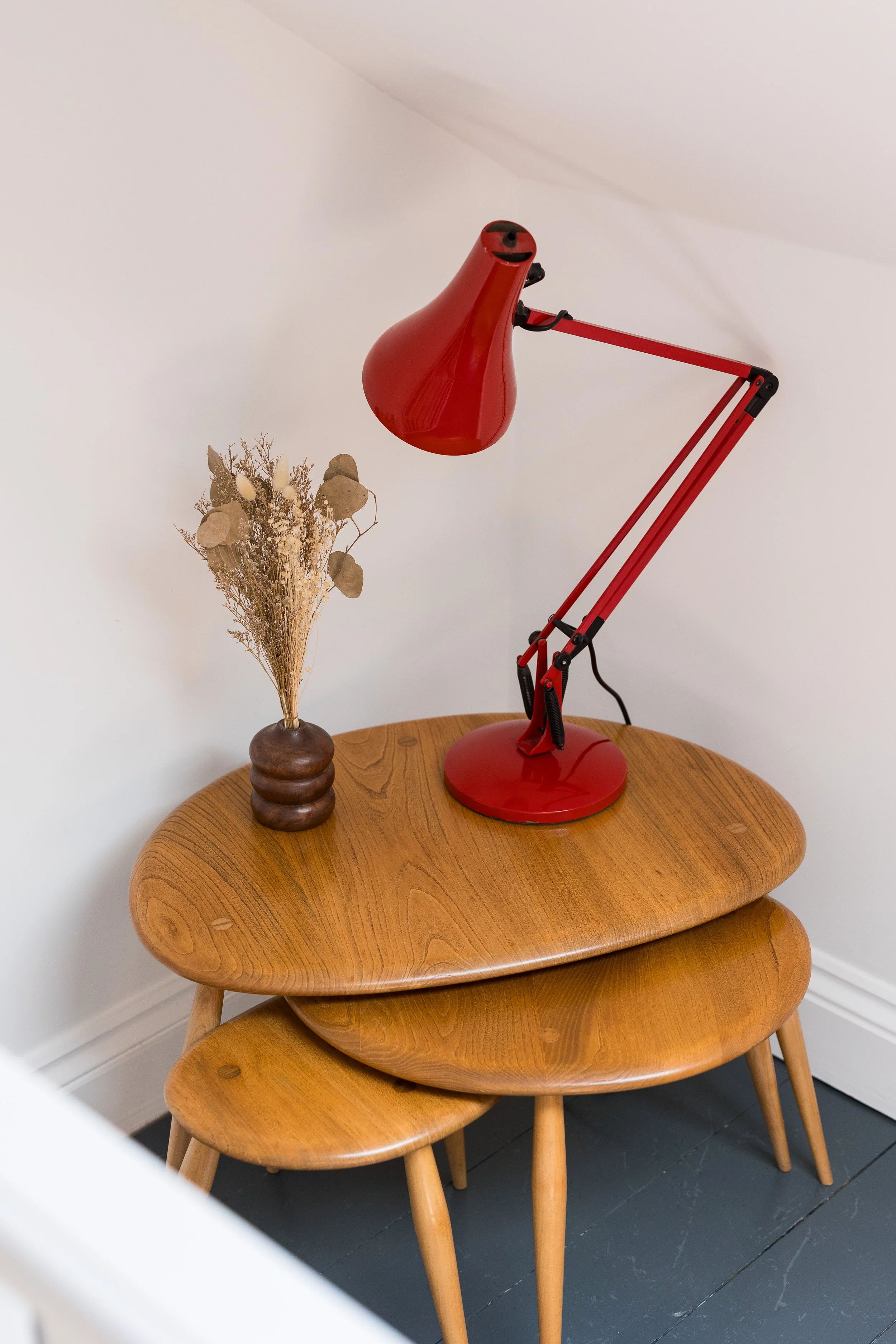 A wooden side table with a small, dark vase holding dried flowers and a tall, red desk lamp with adjustable arm standing on top.