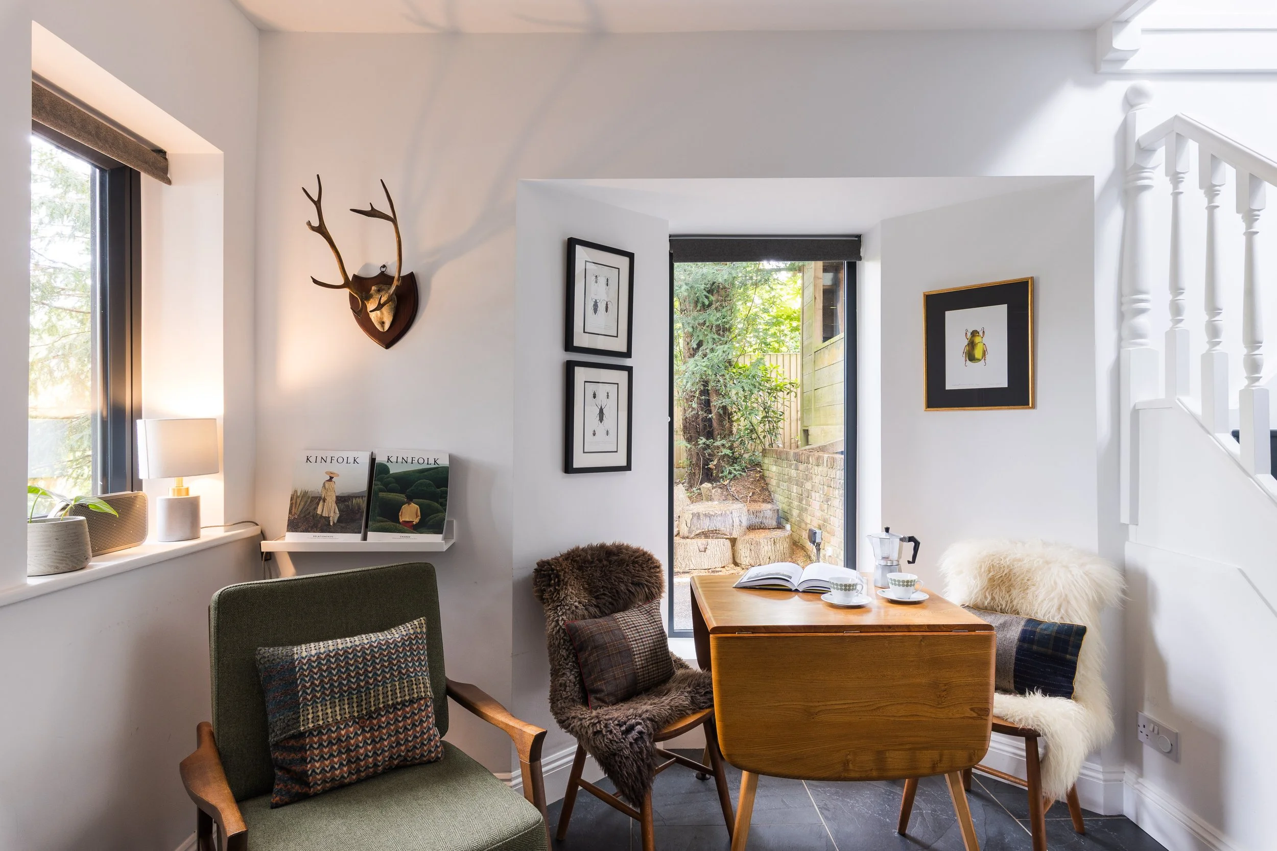 A cozy interior corner with two chairs, one with a multicolored cushion and another with a faux fur throw and plaid cushion. A small wooden table holds an open book and two cups. The wall has framed insect art and a mounted deer head with antlers. Th