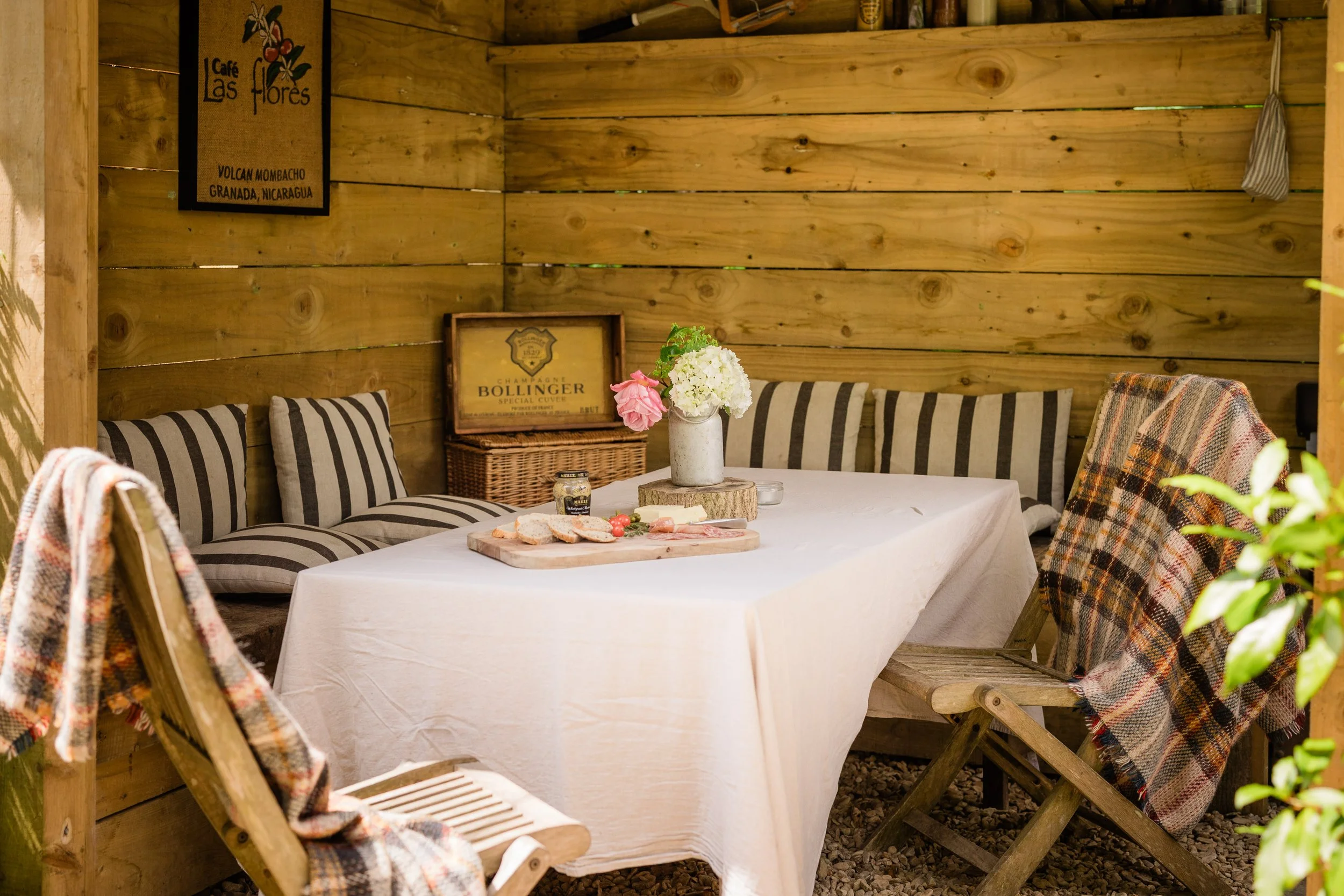 A cozy wooden dining nook with a white tablecloth-covered table, surrounded by striped cushioned chairs and plaid blankets, with a vase of white and pink flowers, and a charcuterie board with cheese, crackers, and tomatoes. The wooden wall has vintag