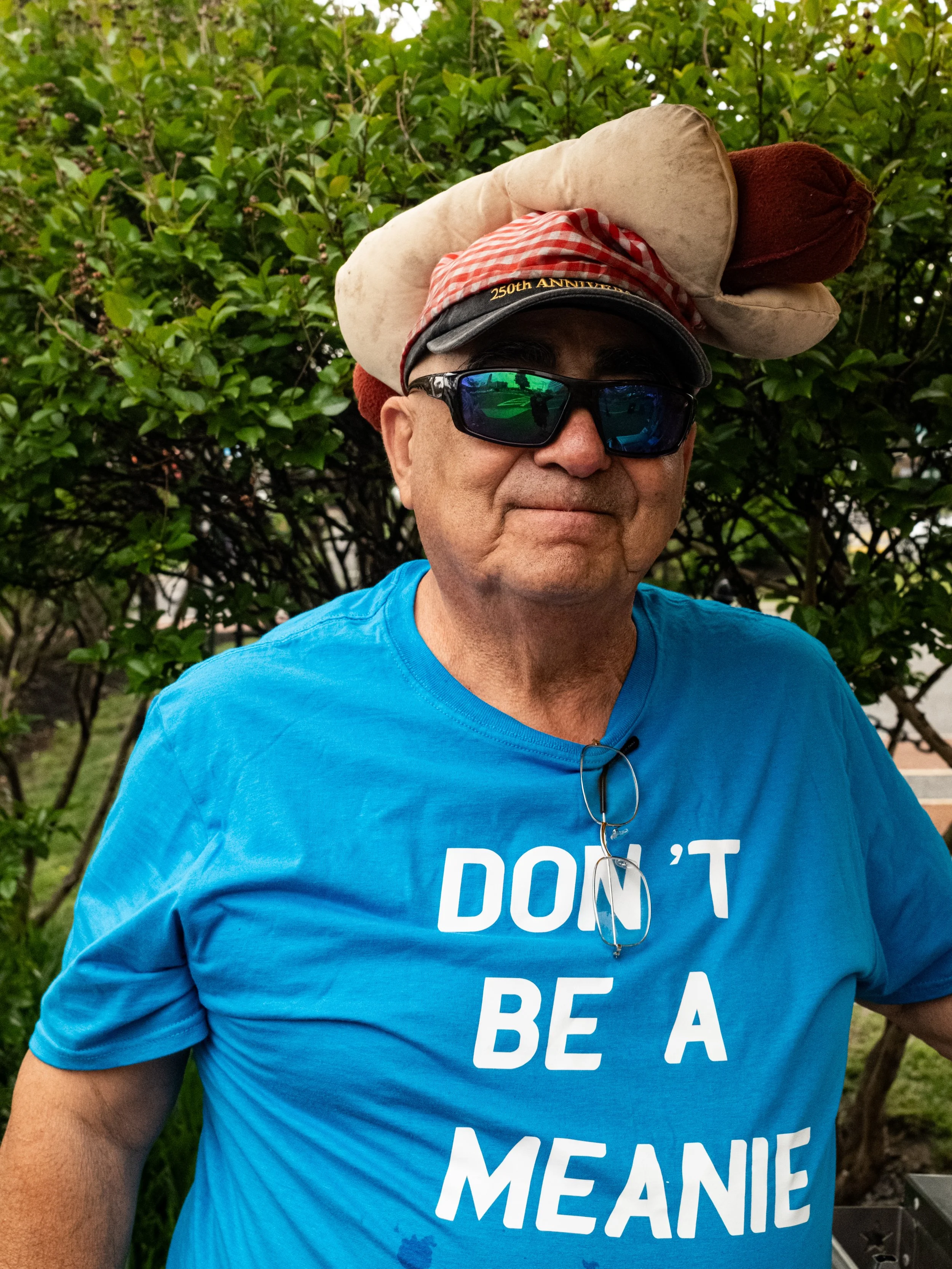 A man wearing a blue T-shirt with white text, a black cap with '250th Anniversary' on it, sunglasses, and a large beige and red plush turkey hat. He is outdoors with green foliage in the background.