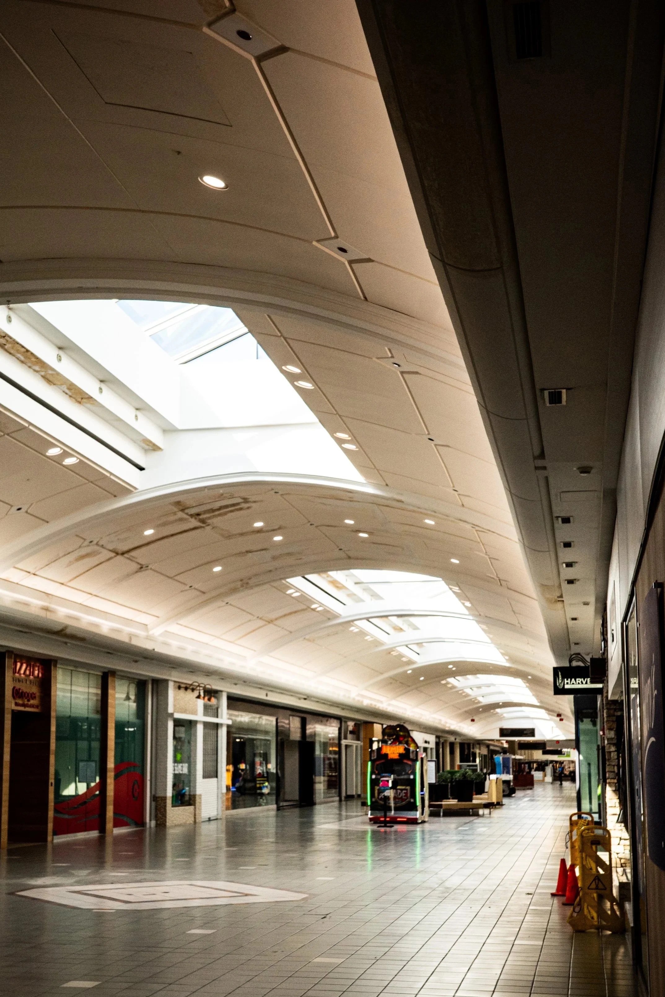 An empty shopping mall corridor with closed stores and overhead lighting, with a ceiling skylight allowing natural light into the space.