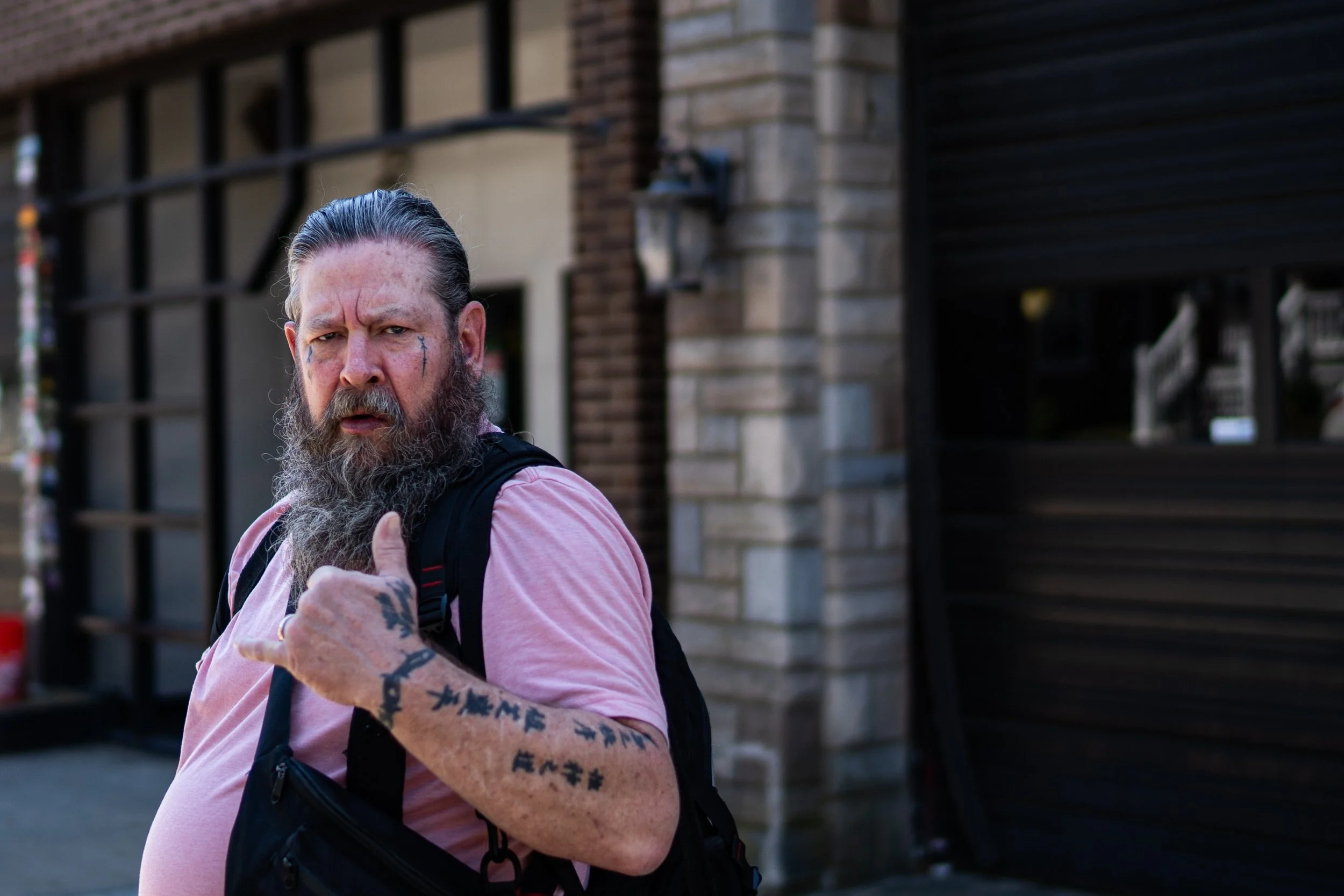 A bearded man with tattoos on his arms and face, wearing a pink shirt and black backpack, giving a thumbs-up gesture in front of a building with a brick and stone facade.
