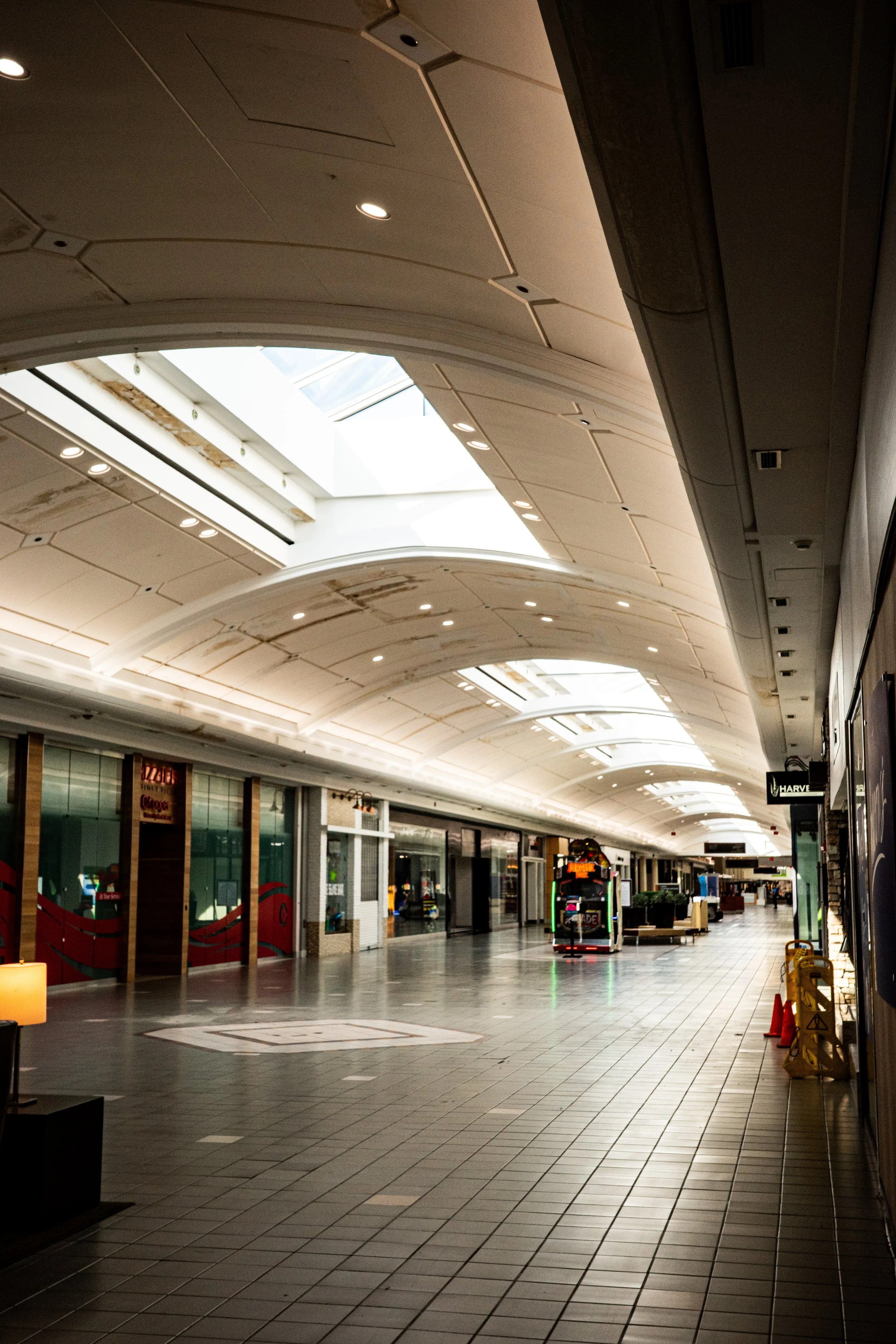 An empty shopping mall corridor with closed stores and overhead lighting, with a ceiling skylight allowing natural light into the space.