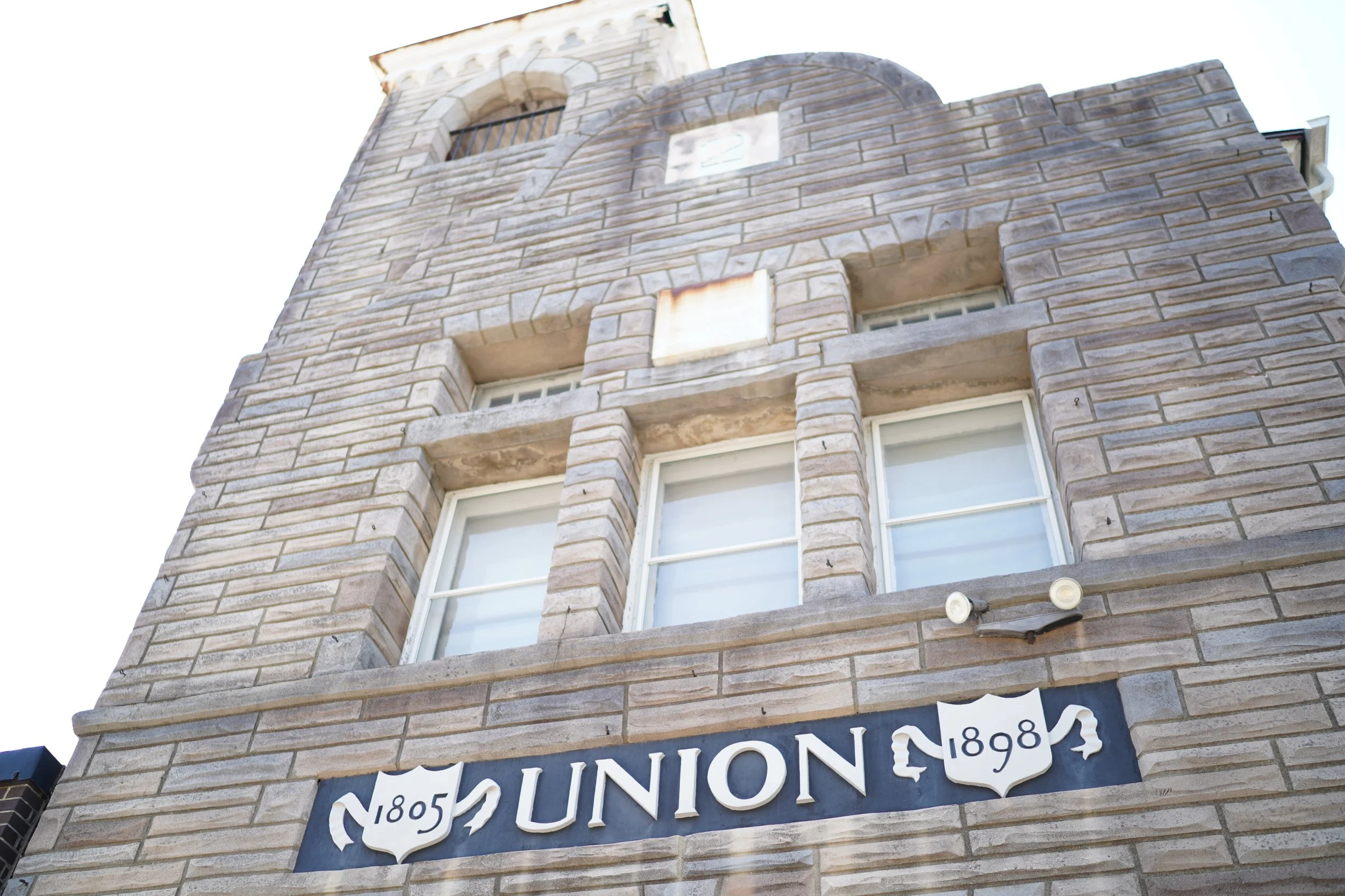 Close-up of a brick building with a sign that reads "Union 1895." The building has two windows, one with bars, and black circular lights mounted below the sign.