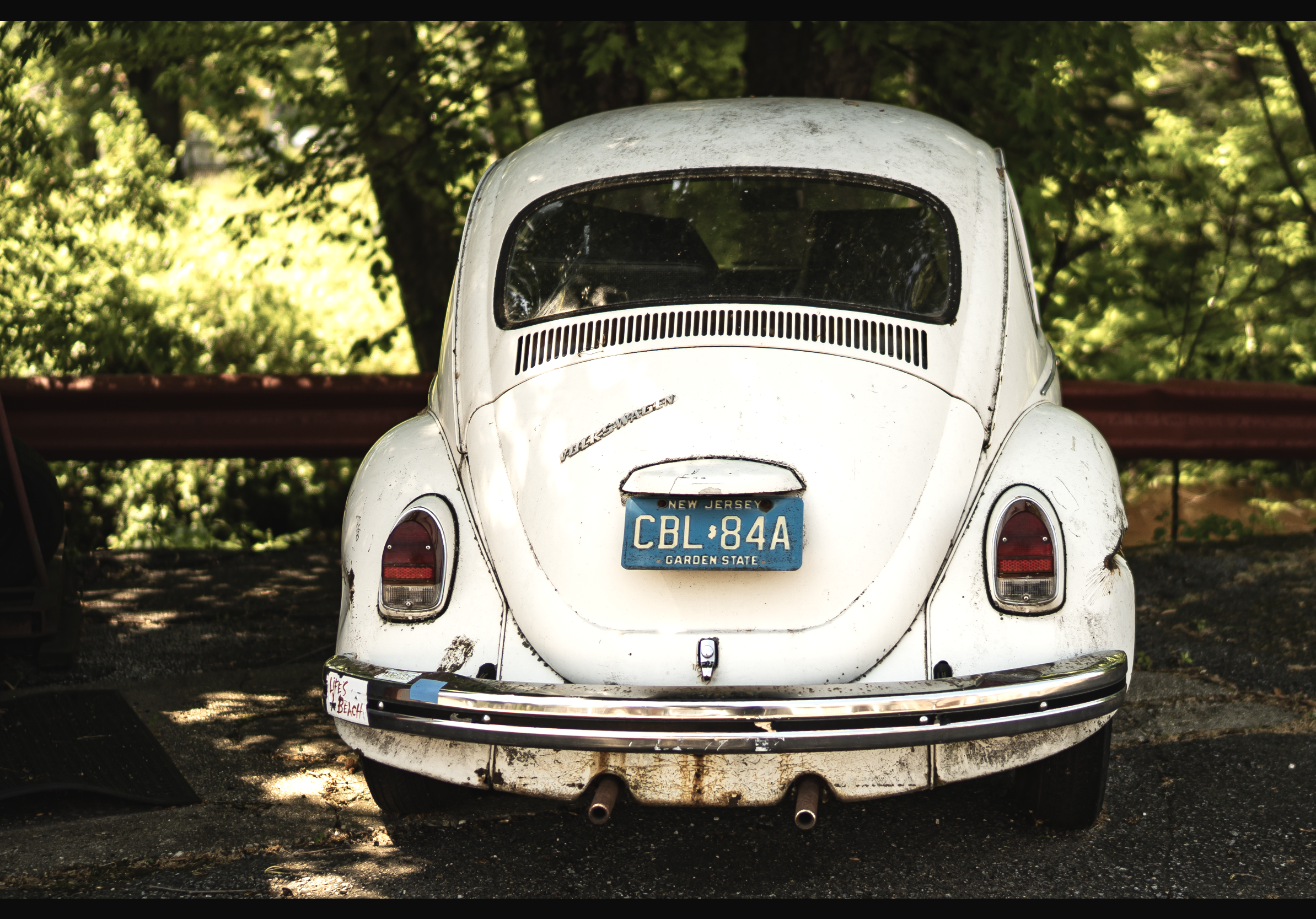 Back of a vintage white Volkswagen Beetle with a blue New Jersey license plate, parked outdoors among trees.