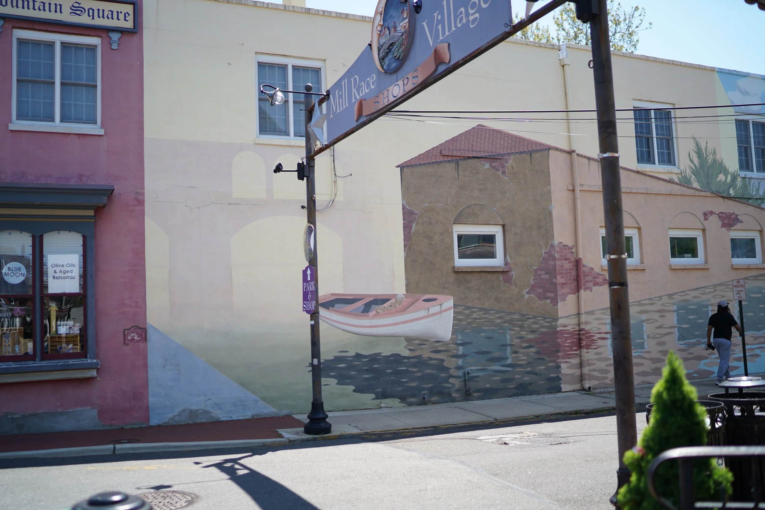 Street view showing a corner with colorful painted building walls, a sign that reads 'Mill Race Shops,' and a person walking on the sidewalk. There are storefront windows, a decorative boat replica on a pole, and some street signs and plants in the f