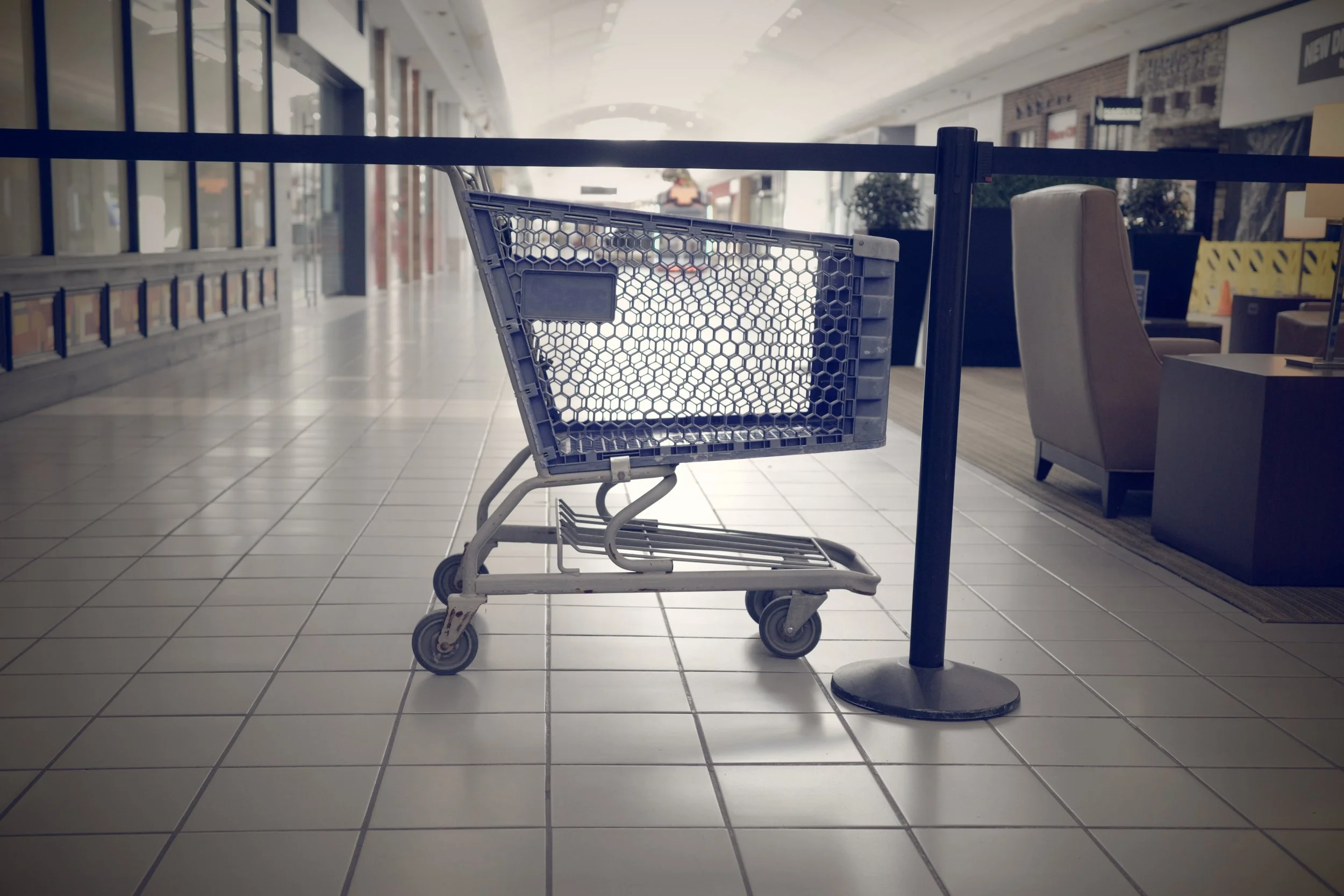 An empty shopping cart in an indoor shopping mall, placed in front of a black pole and a beige armchair, with storefronts and tiles in the background.