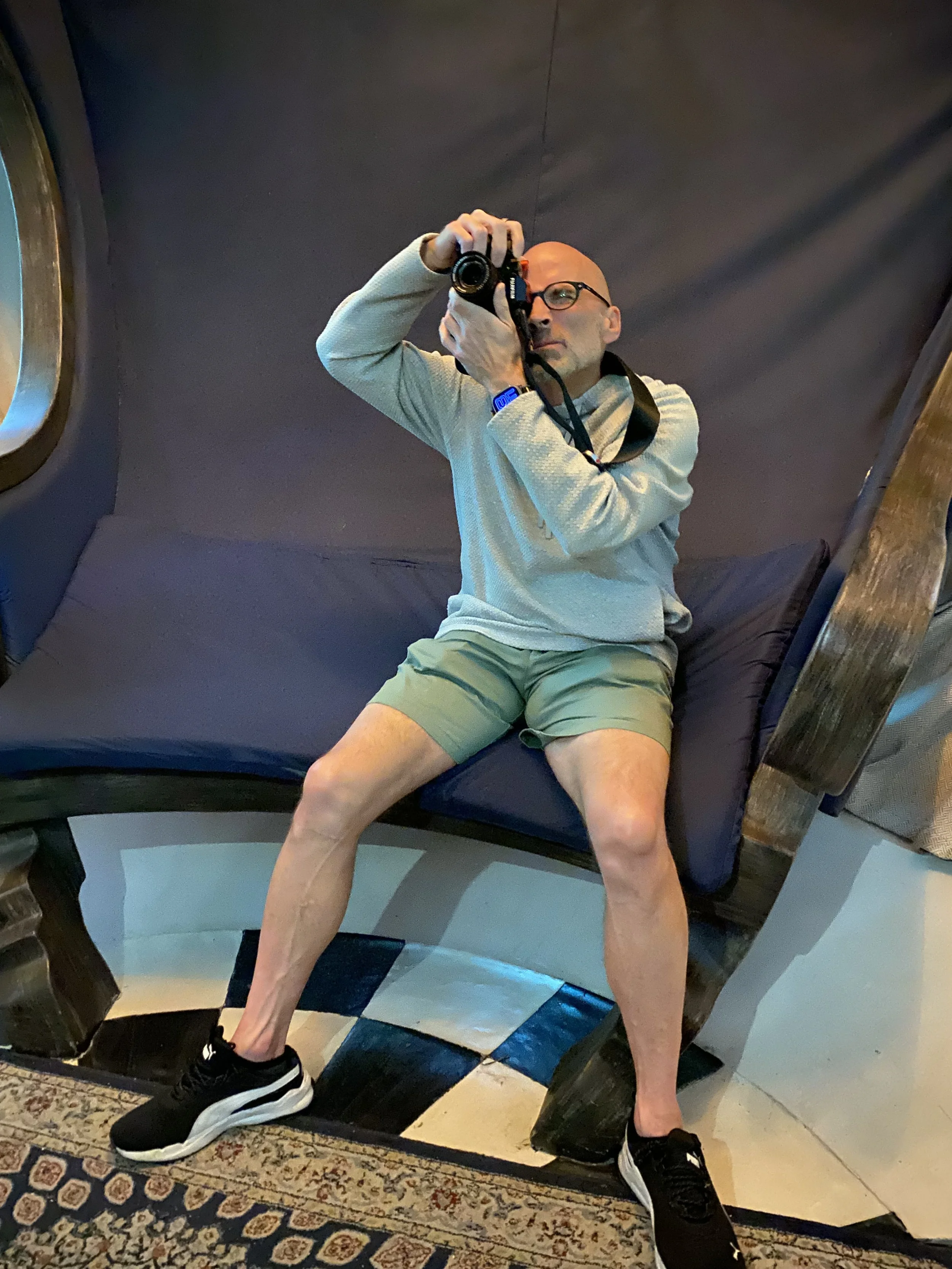 A man taking a mirror selfie in a room with dark walls, sitting on a curved bench, wearing glasses, a light-colored hoodie, khaki shorts, and black sneakers, with one foot on the floor and the other slightly raised.