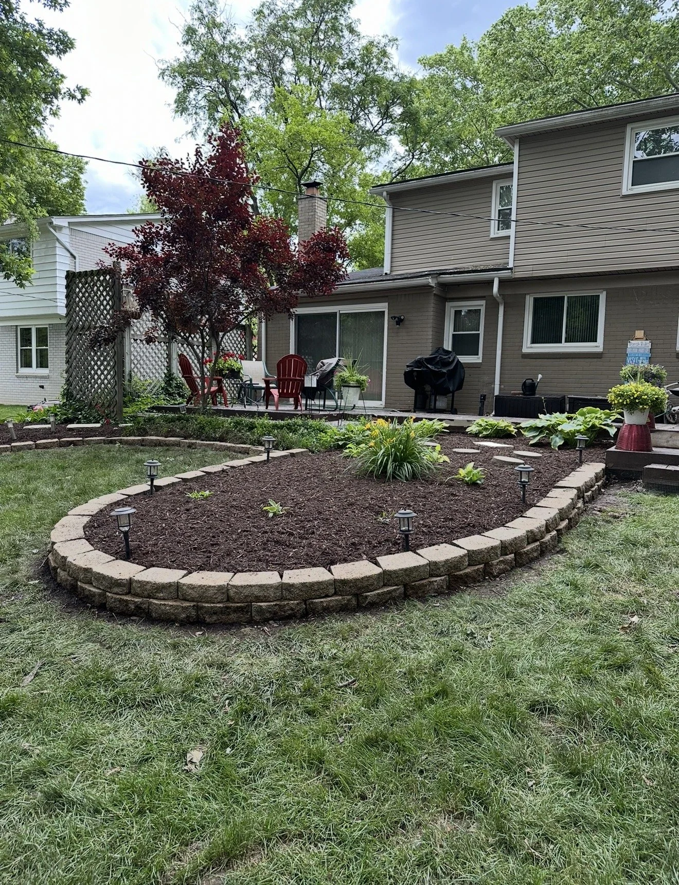 A backyard garden with a raised flower bed surrounded by brick edging, featuring small plants and garden lights. Behind the garden, there is a seating area with outdoor chairs and a table, and a patio with a barbecue grill. The house in the background has a brown exterior with multiple windows, and is surrounded by green trees.