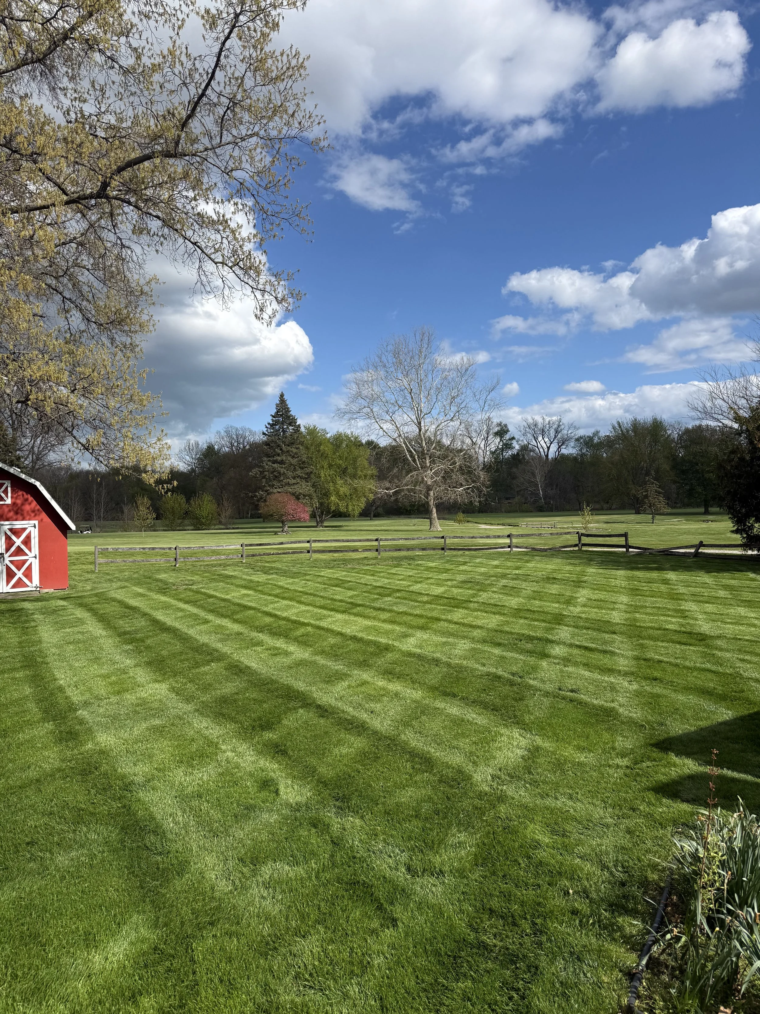 A scenic view of a well-maintained grassy field with a red barn on the left side, a white split-rail fence, and a variety of trees in spring bloom under a partly cloudy sky.