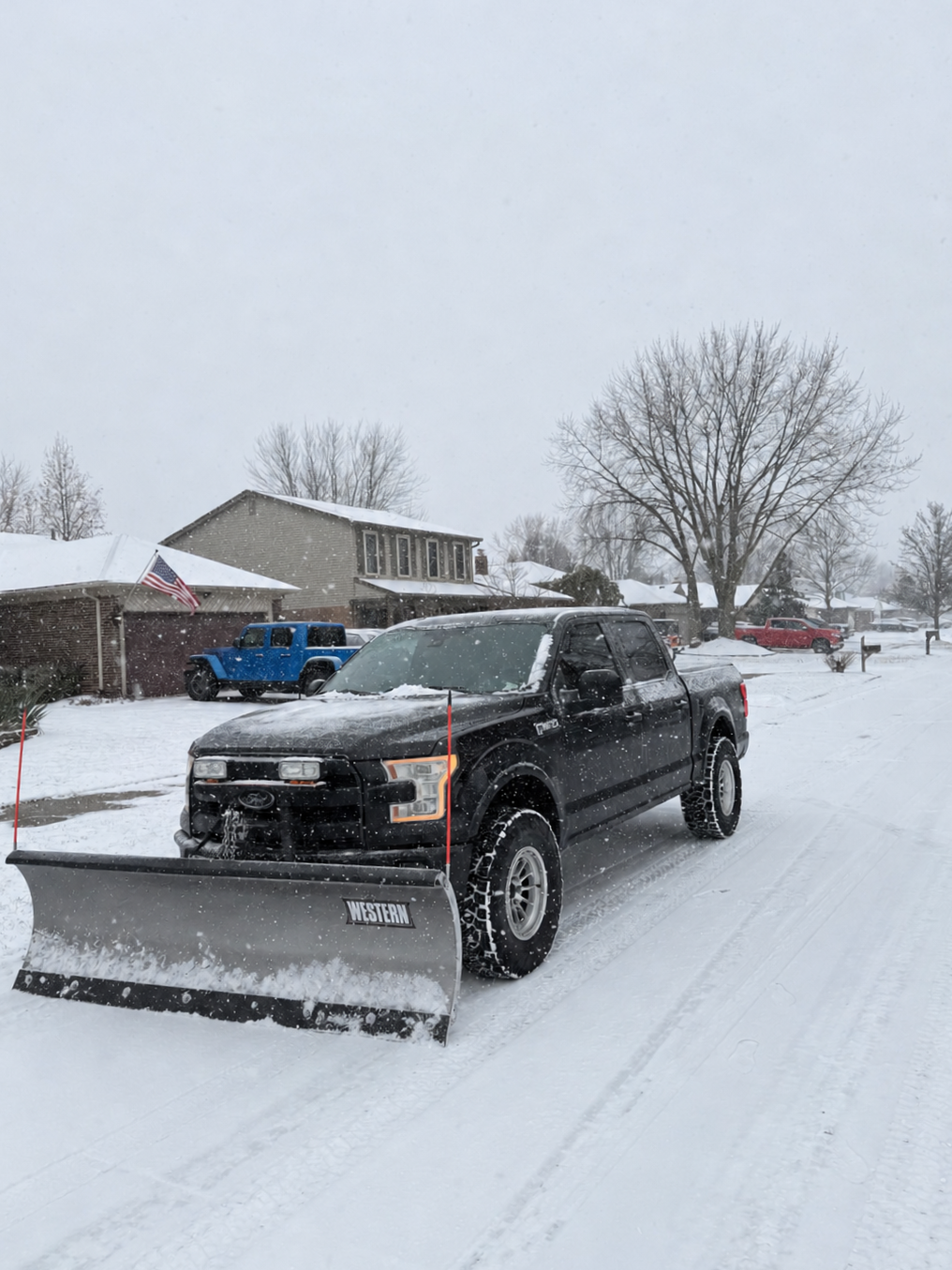 Black pickup truck with a snow plow attached, clearing snow on a residential street during snowfall in winter.