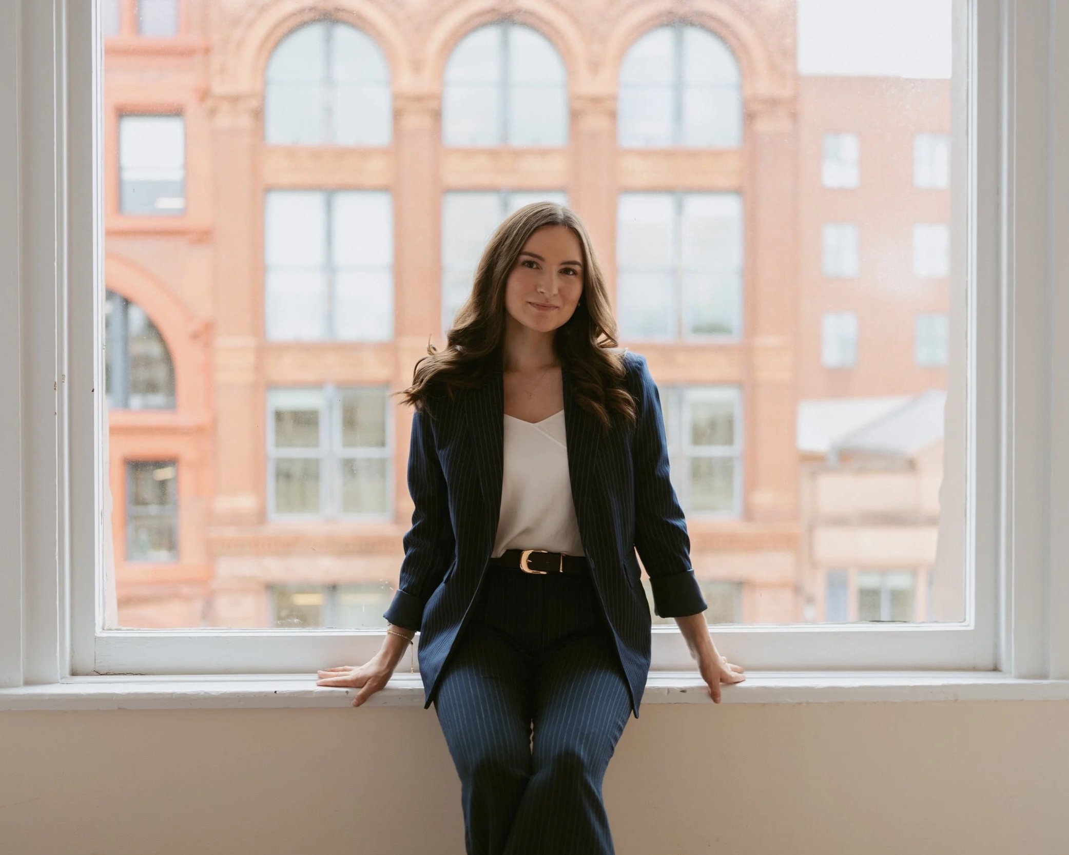 A woman in a navy-blue pinstripe blazer and matching trousers standing by a large window with a brick building outside, smiling softly.