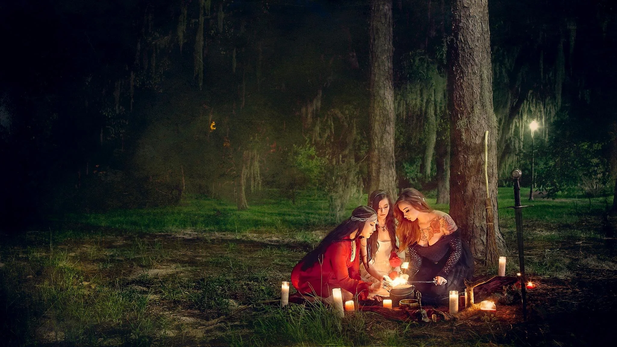 Three women gathered around candles on the ground in a forest at night, looking down into a cauldron, with trees and streetlights in the background.