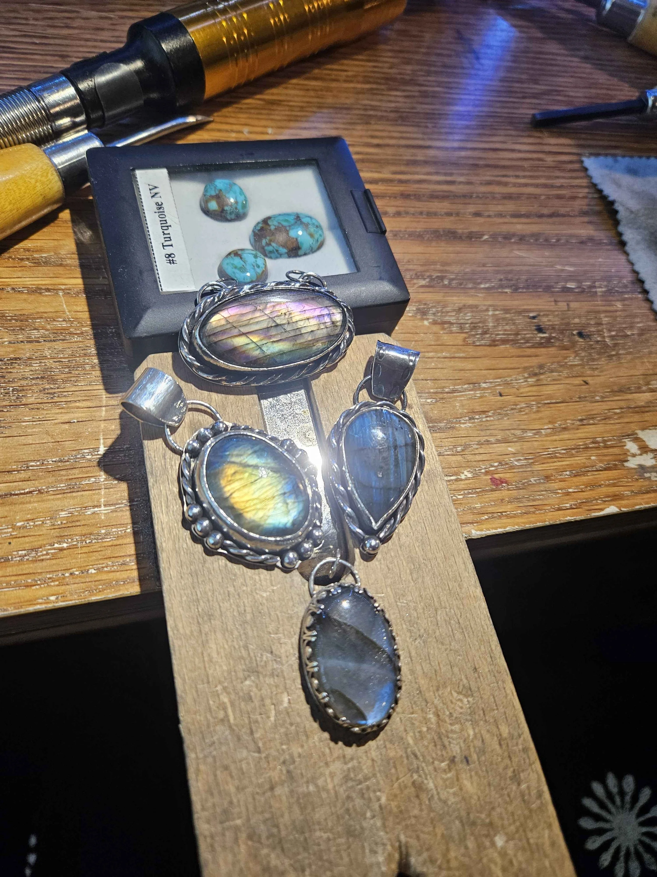 A display of turquoise and labradorite jewelry on a wooden surface, including bracelets and a large pendant with colorful iridescent stone, along with a box containing polished turquoise stones. A jeweler's tool and a polishing cloth are also visible.